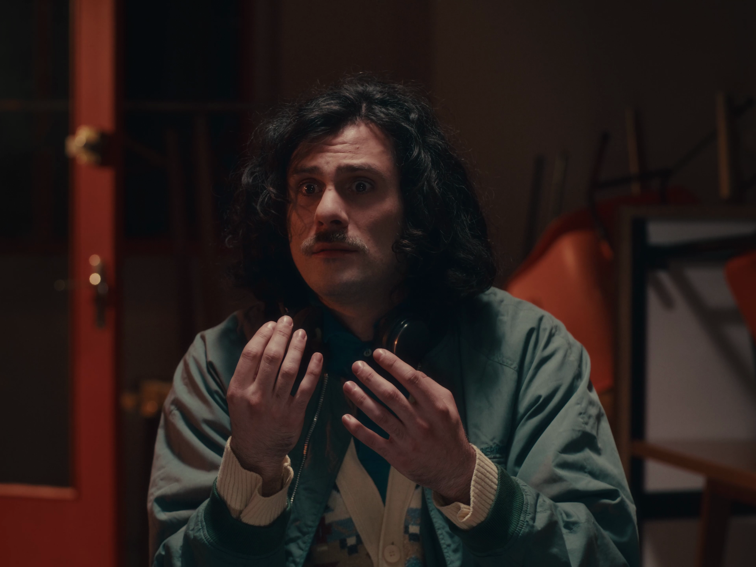 Un homme aux cheveux longs et bouclés, portant un blouson en nylon et tenant un casque audio, se trouve dans une pièce sombre avec des étagères et des chaises empilées en arrière-plan.