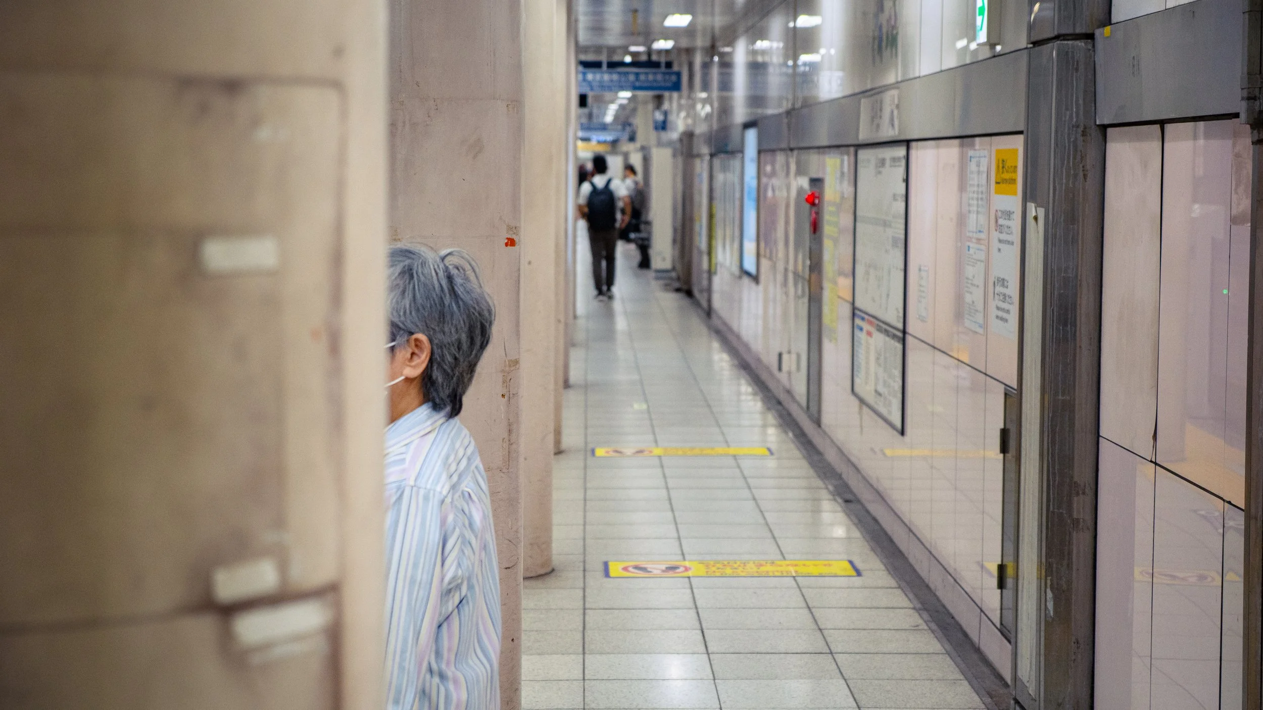 Une personne âgée portant un masque, regardant depuis un coin d'un couloir de métro ou de gare, avec d'autres personnes en arrière-plan en marche.