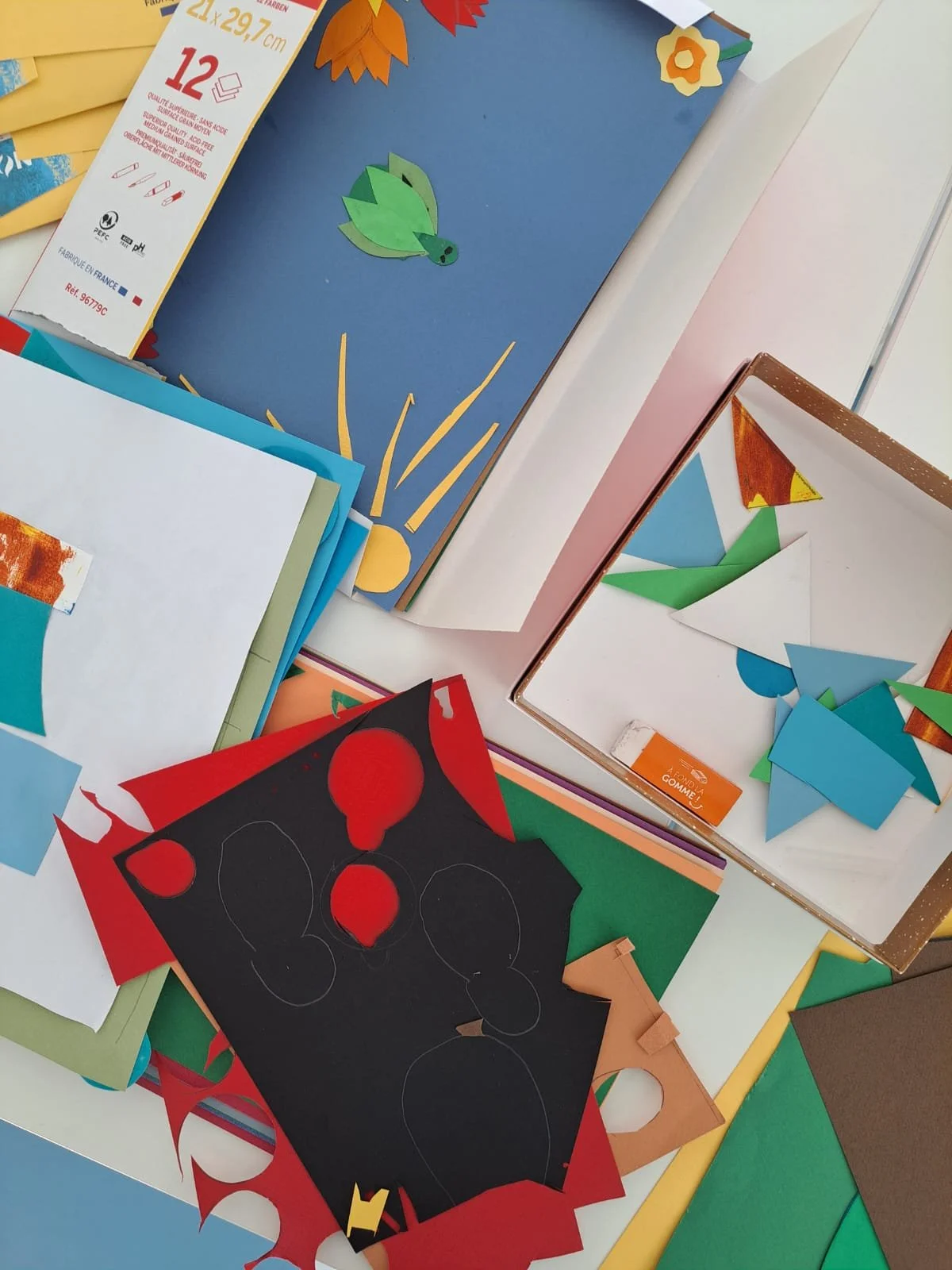 Une table avec divers papiers colorés, des formes découpées, un cadre en carton et des feuilles pour activités DIY en Guadeloupe.
