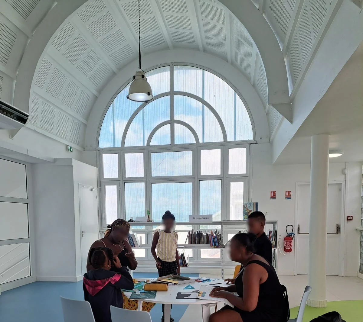 Groupe de personnes, incluant des enfants et des adultes, rassemblées autour d'une table dans un espace lumineux, en train de discuter ou de réaliser un atelier d'art plastique, coloriage, papiers découpés et collage.
