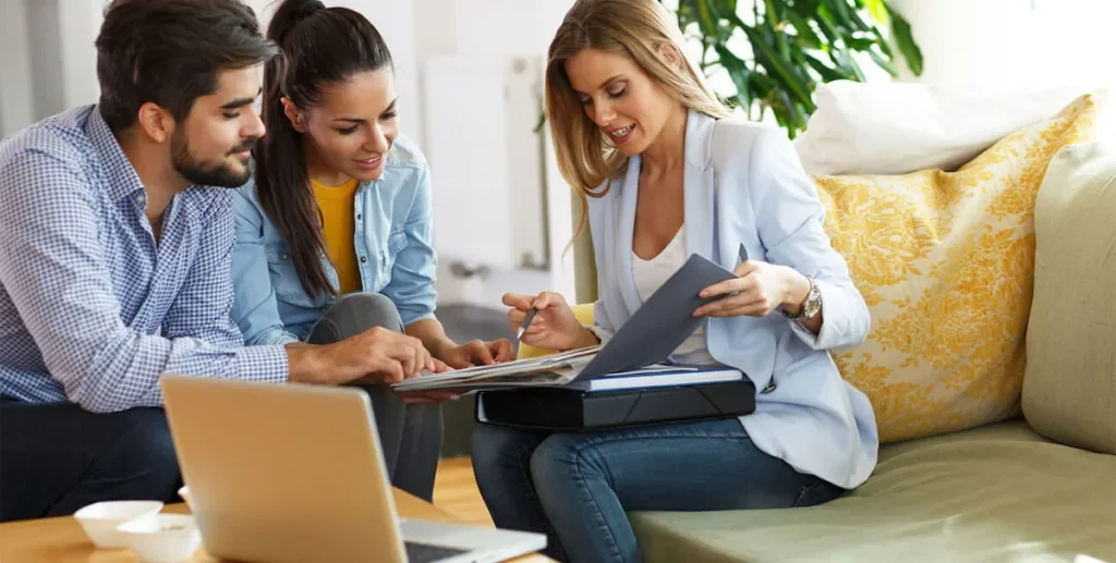 A woman in a light blazer shows a document to a couple sitting on a sofa, helping them with their health insurance needs.