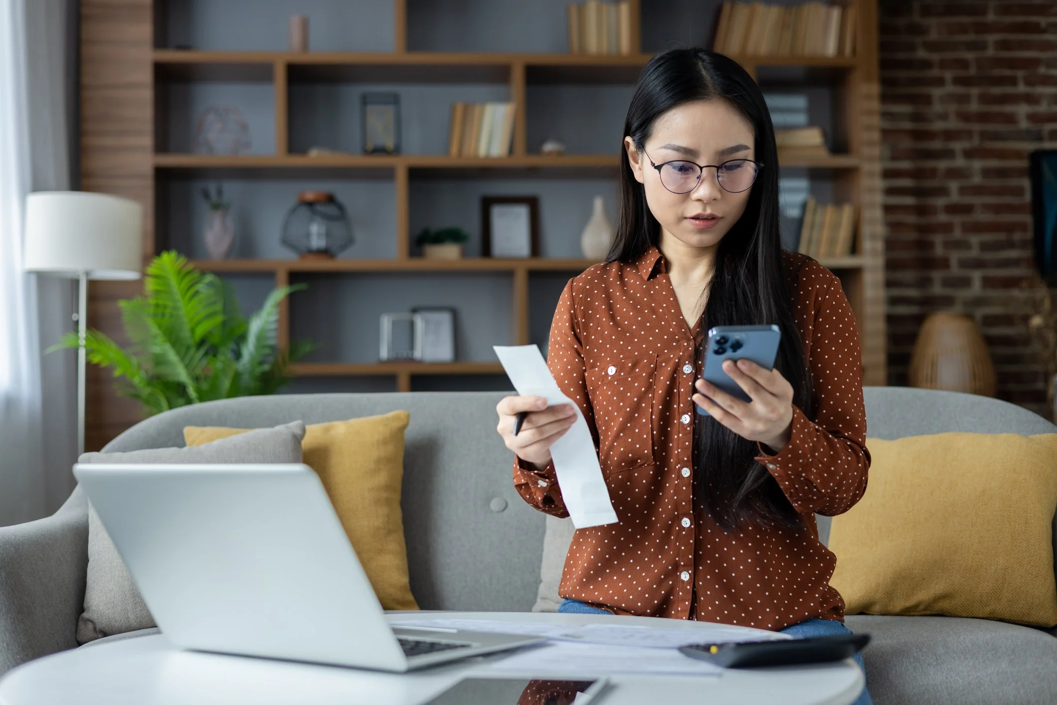 A woman with glasses and long dark hair looking at her phone while holding a receipt, sitting on a sofa in a living room with a laptop and papers on a coffee table.