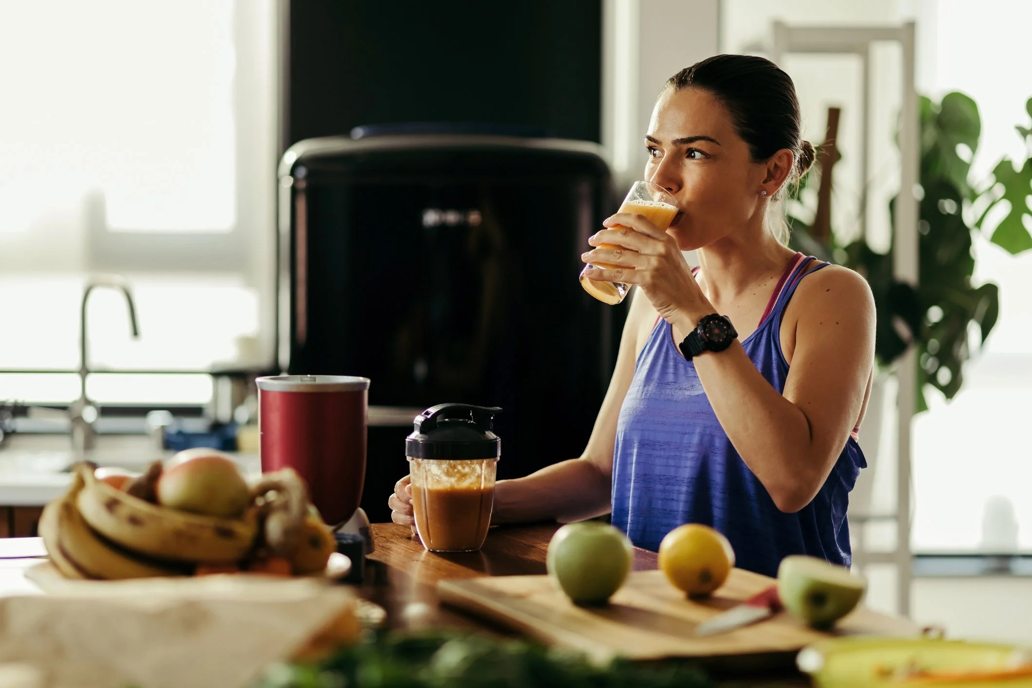 Woman in athletic wear drinking from a glass of juice in a kitchen with fruits, a blender, and a cutting board with apples and a knife on the counter.