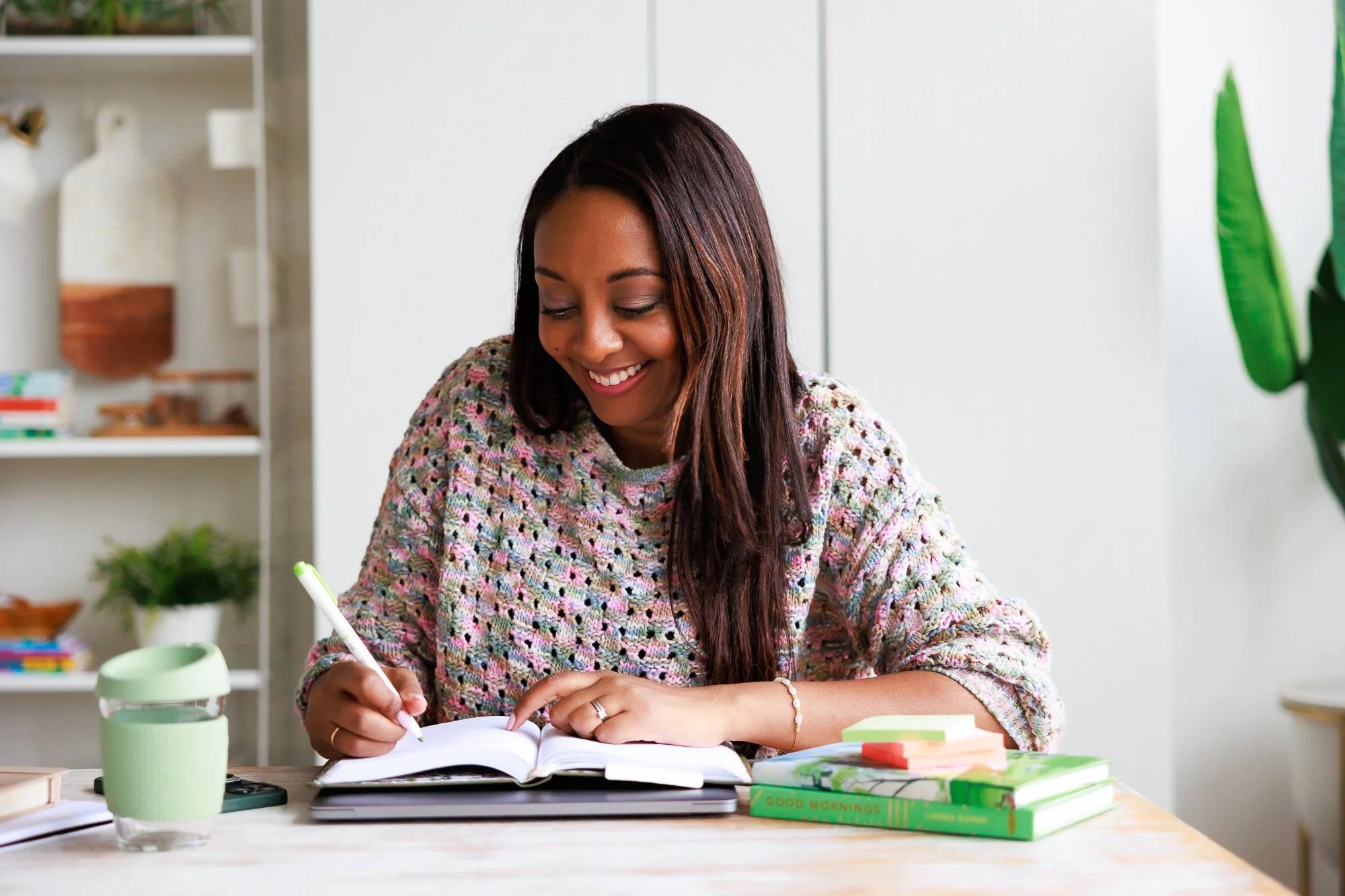 A woman with long dark hair smiling while writing in a notebook at a desk with books, sticky notes, a green mug, and a plant in the background.