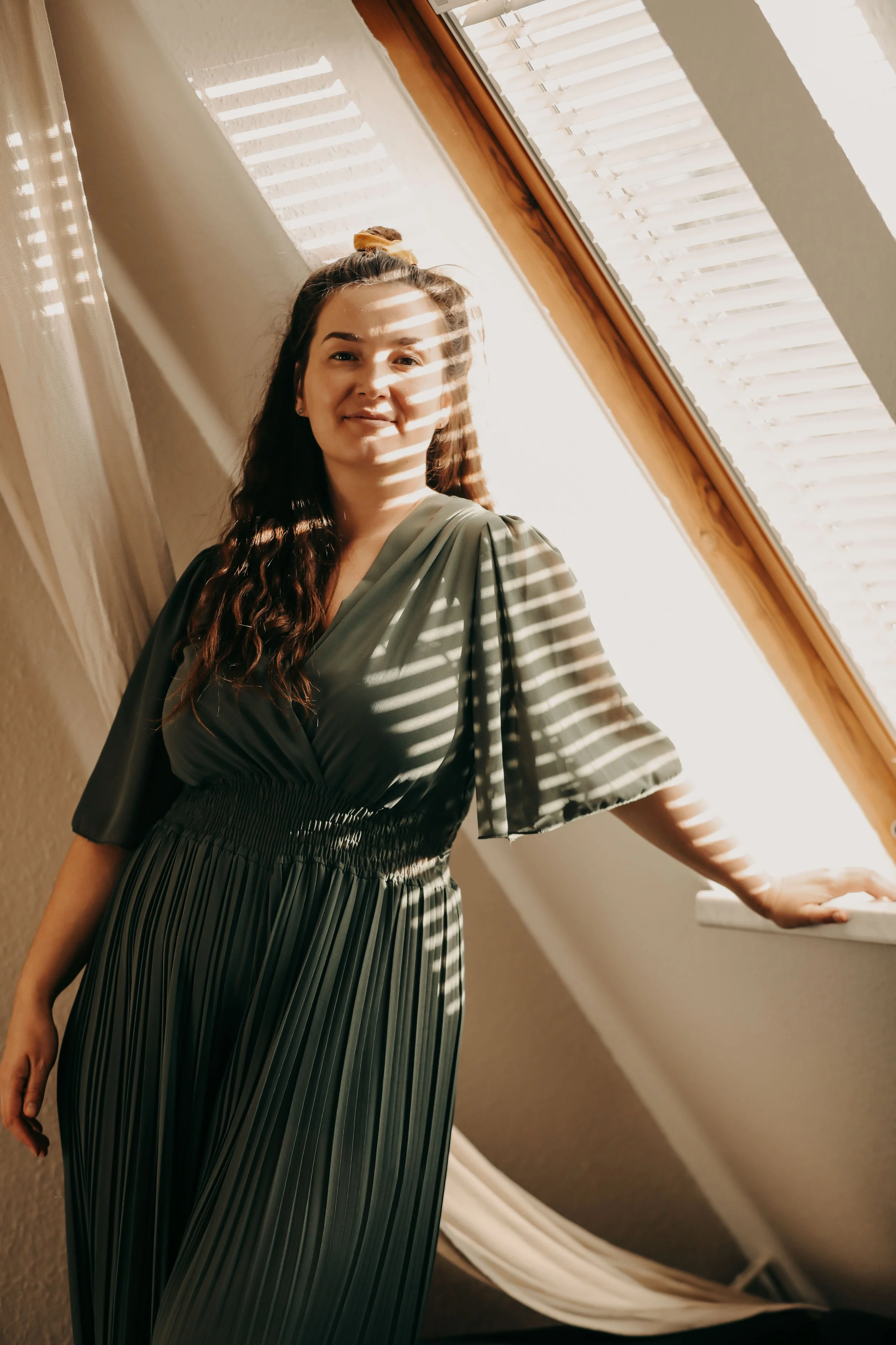 Christin Reese – Energiearbeit – in einem grünen Jumpsuite steht vor einem Fenster mit Jalousien, durch die Sonnenlicht in Streifen fällt, im Raum mit gedämpften Farben.