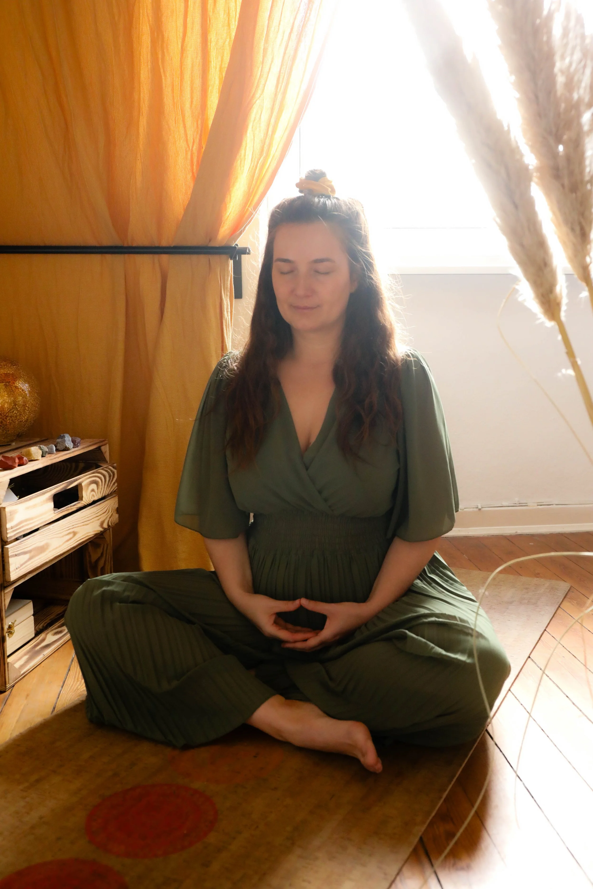 Frau sitzt kreuzleg auf einem Teppichboden mit gekreuzten Beinen, Hände in Meditationshaltung, im stillen Raum mit Sonnenlicht durch das Fenster, umgeben von Pflanzen und Dekorationen.