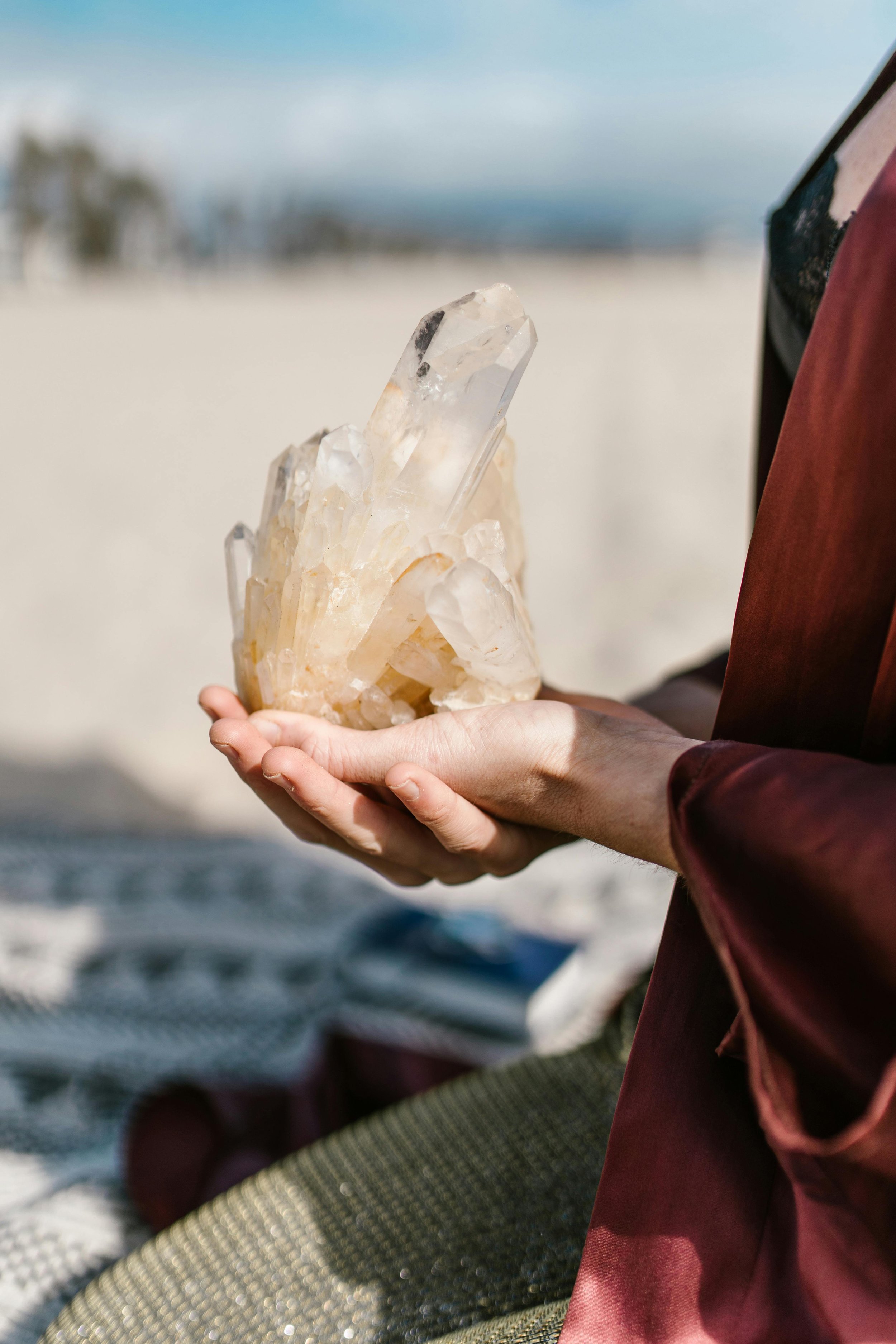 Hand hält einen großen Kristall mit anderen Kristallen im Hintergrund, am Strand, bei Sonnenlicht.