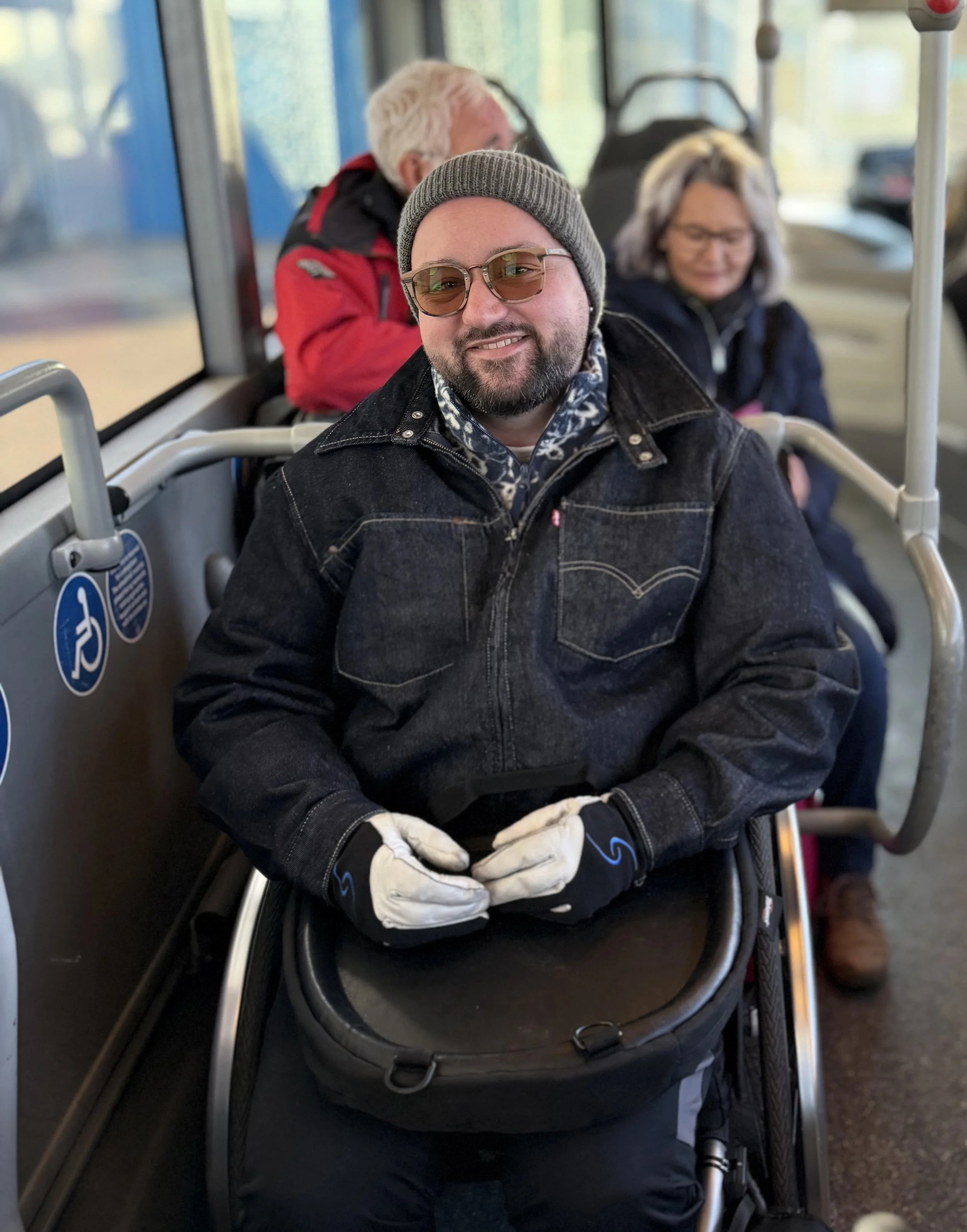 Alex Clare-Young in an accessible space on a bus. Alex is a white man with tinted brown glasses and a black and grey beard. They are wearing a blue denim jacket and a blue and white fleece as well as a beanie hat. They are sitting in a wheelchair.
