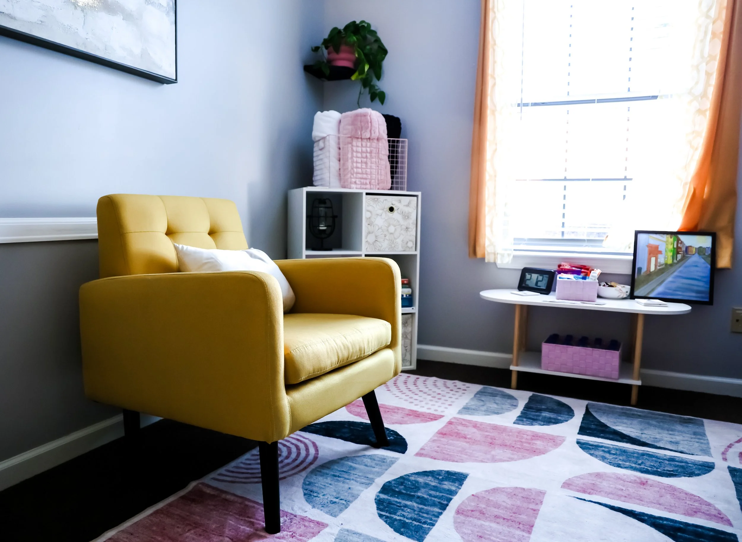 A bright living room with a yellow armchair, a white side table, a window with orange curtains, and a colorful geometric rug on the floor.