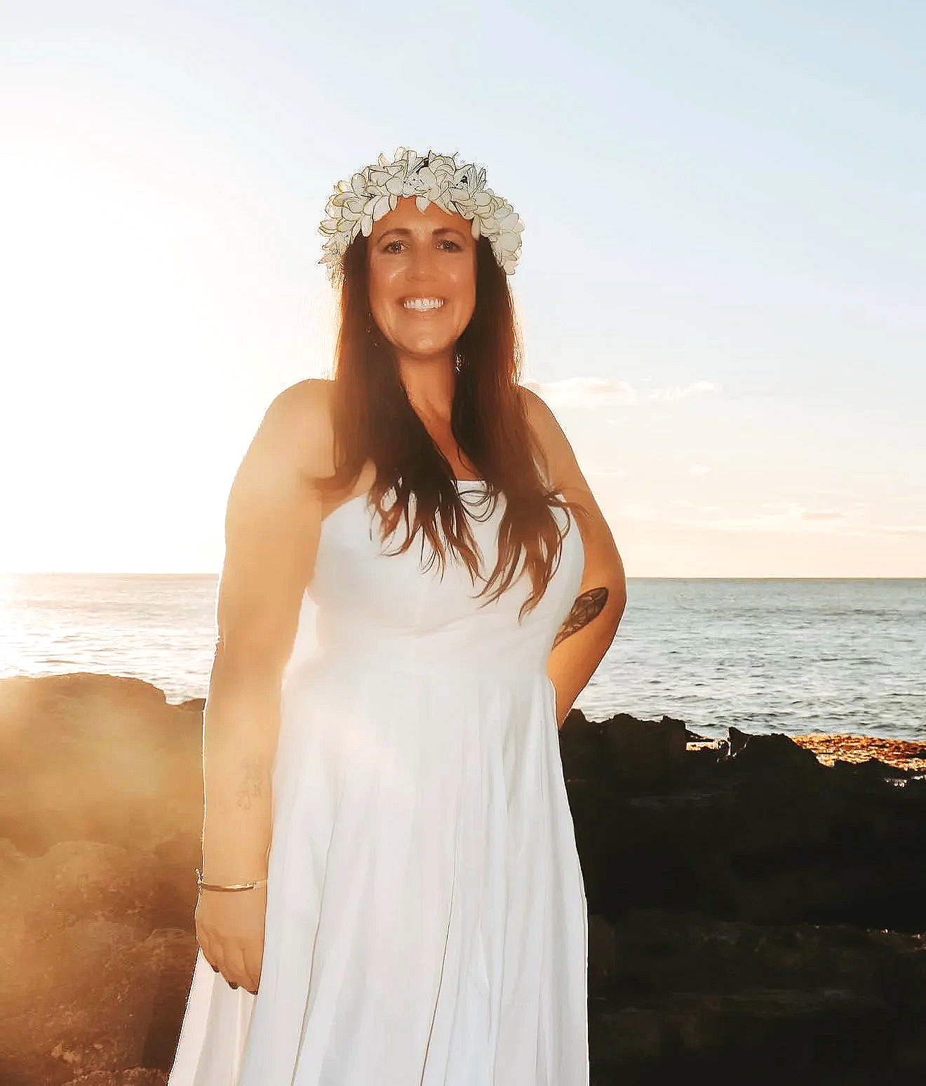 Woman in white dress smiling at the camera on a beach during sunset, wearing a floral crown.