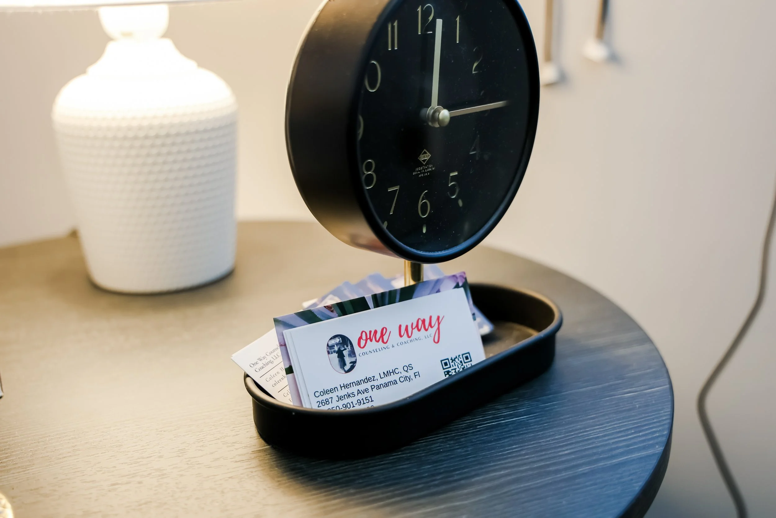 Business cards placed in a black tray beneath a black round clock on a wooden nightstand, with a white textured lamp in the background.