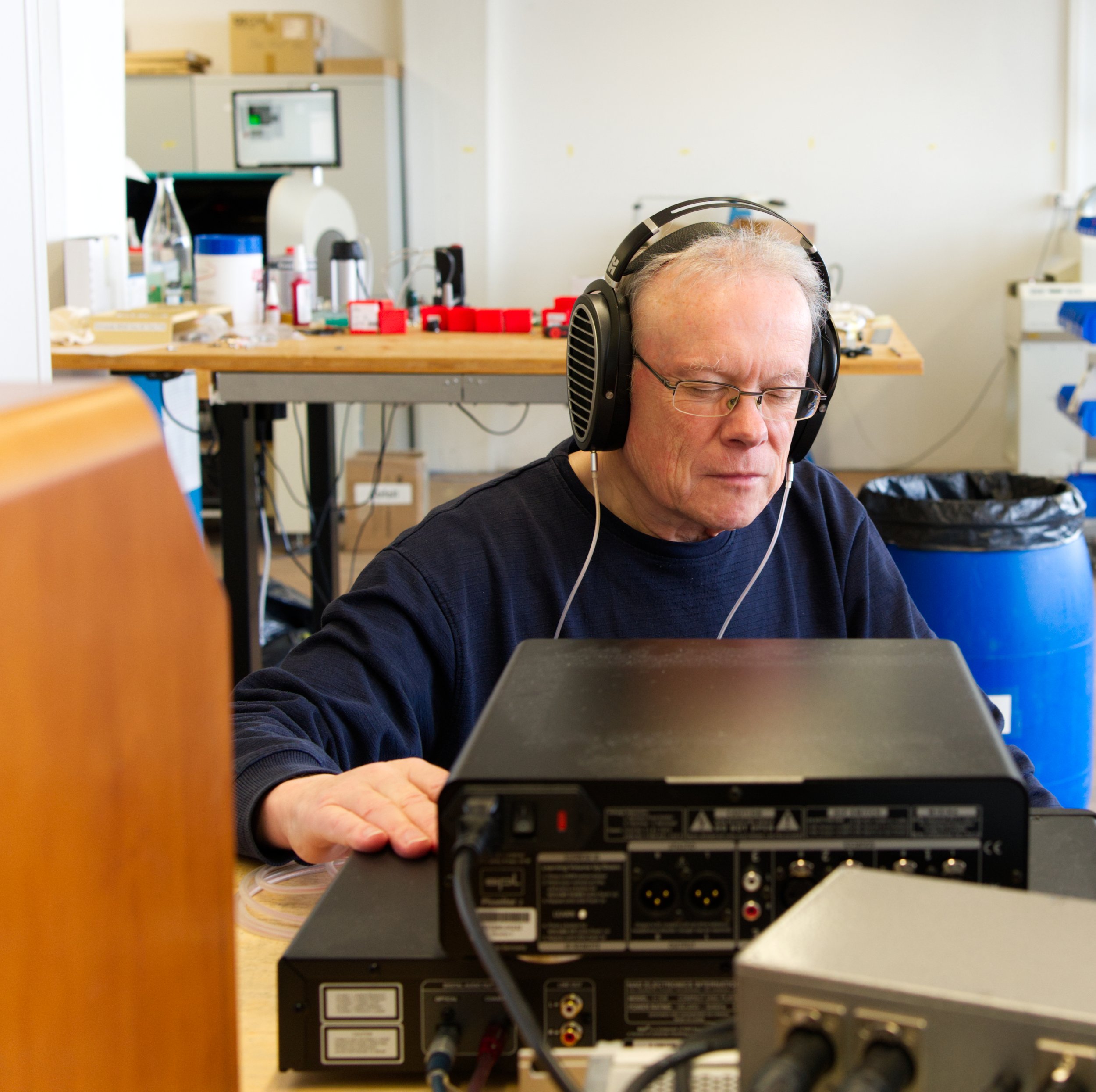 Ein älterer Mann trägt einen schwarzen Pullover, eine Brille und große Kopfhörer, während er an einem Audiogerät arbeitet in einem Werkstatt- oder Laborraum mit einem Arbeitstisch im Hintergrund.