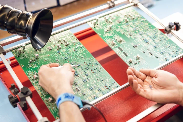 Zwei Personen arbeiten an einer elektronischen Leiterplatte, eine Person benutzt ein Werkzeug, während die andere Hand bereit ist, bei der Montage zu helfen.