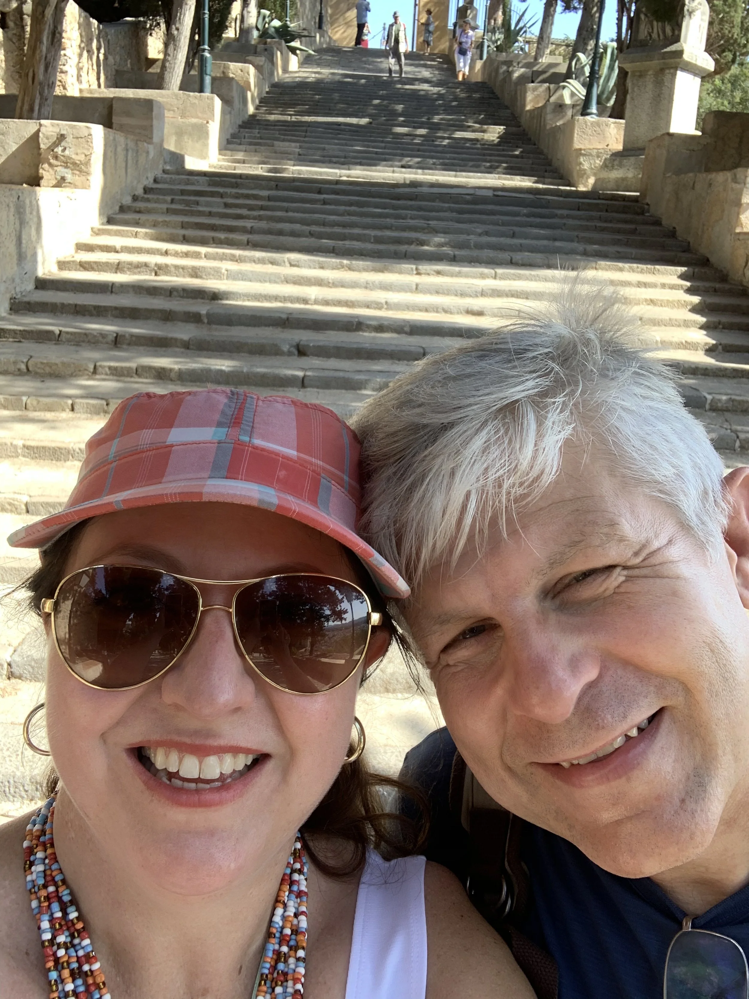 A smiling woman wearing sunglasses, a pink hat, and beaded necklaces, and a smiling man with gray hair, taking a selfie in front of a stone staircase.