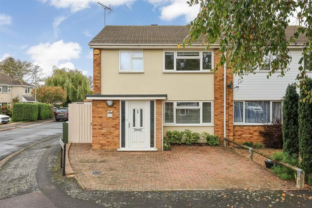 Front view of a two-story house with a white door, brick accents, and beige and white exterior walls. There is a small hedge below the window and a driveway with a brick paving pattern.