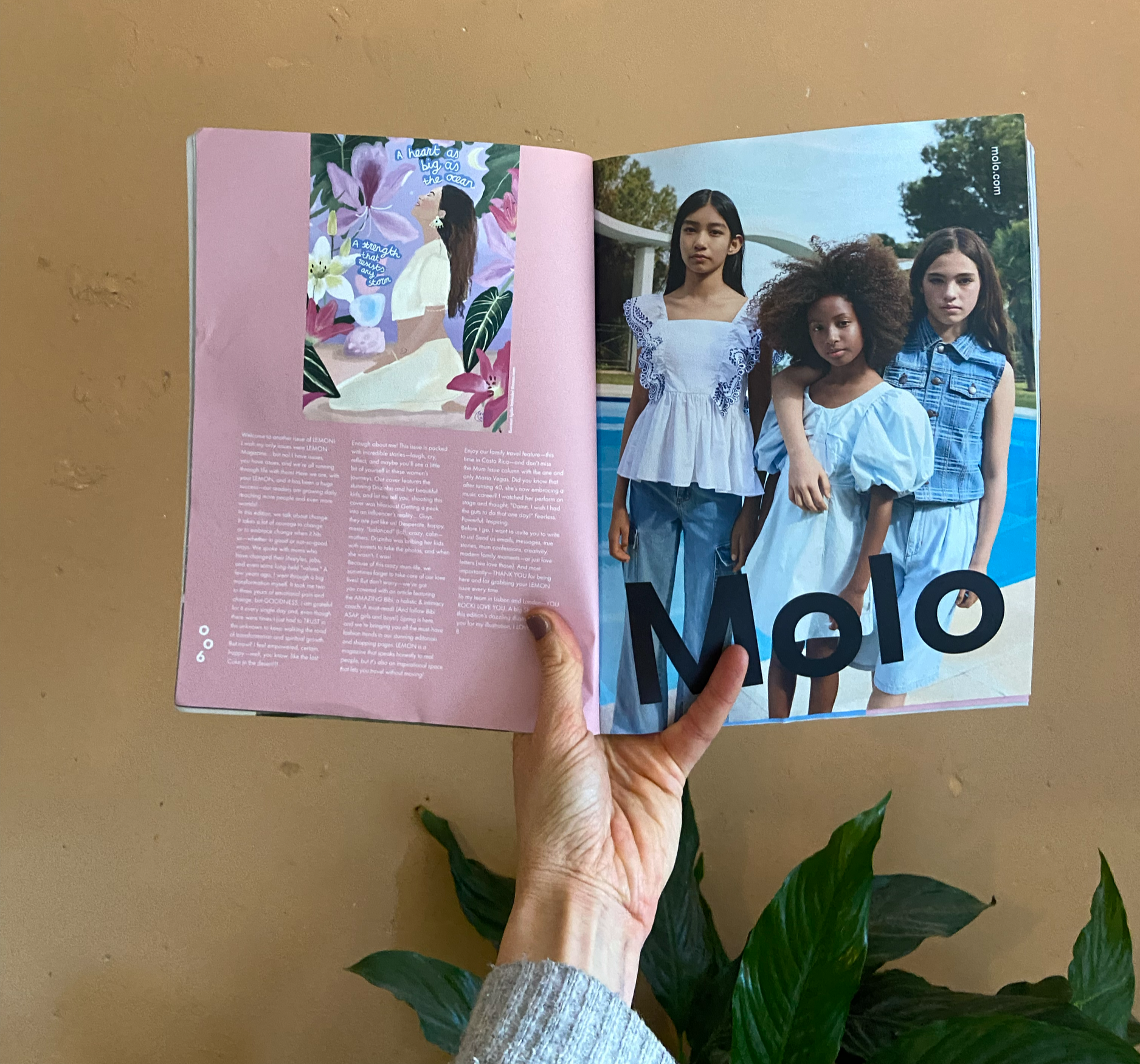A hand holding open a magazine on a beige surface, revealing a two-page spread. The left page features an illustration of a woman with long brown hair among pink and purple flowers with the text "A heart as big as the ocean" and "A strength that caus