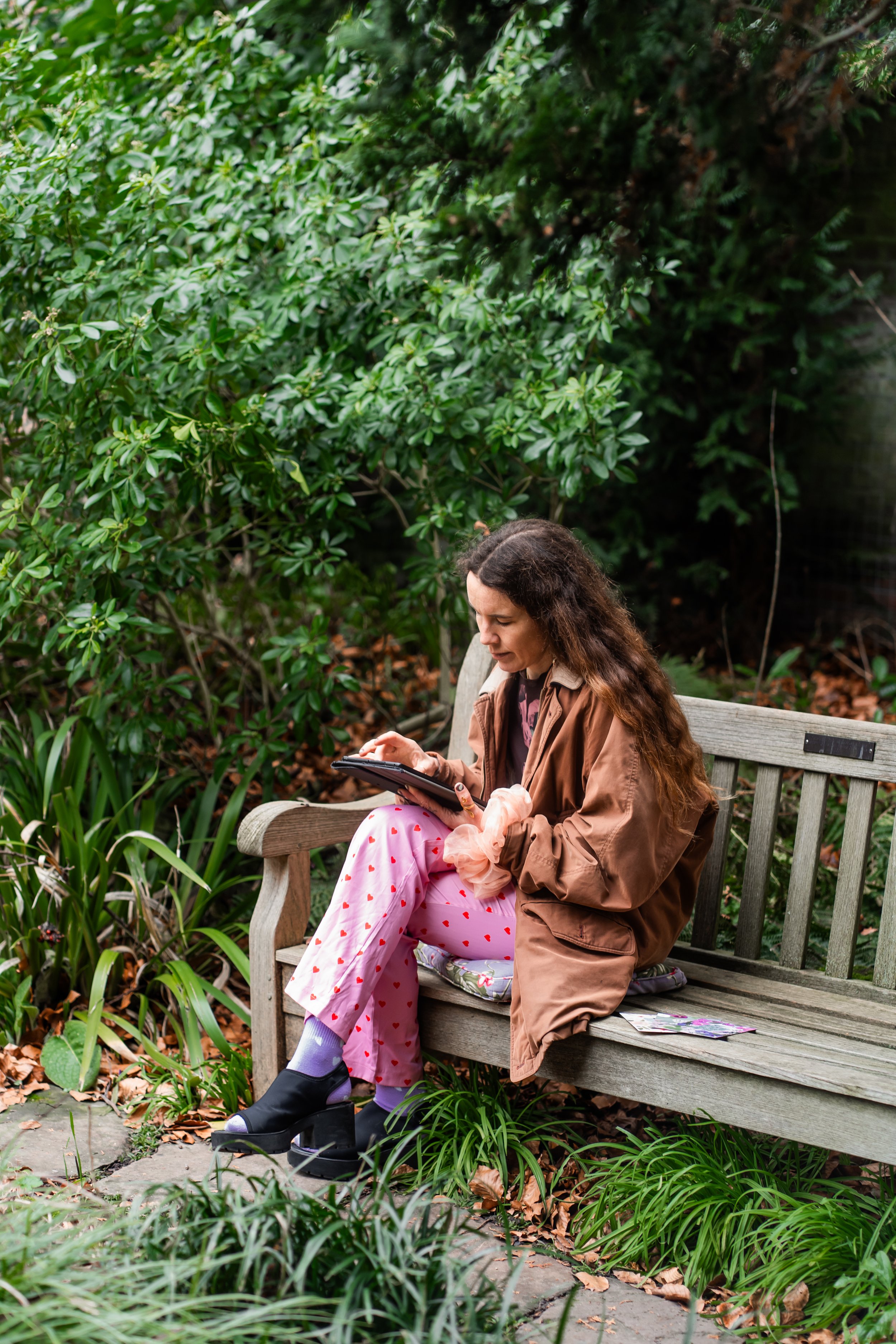 A young woman with long curly hair sitting on a wooden park bench, looking at her phone, surrounded by green bushes and plants, with an overcast sky.
