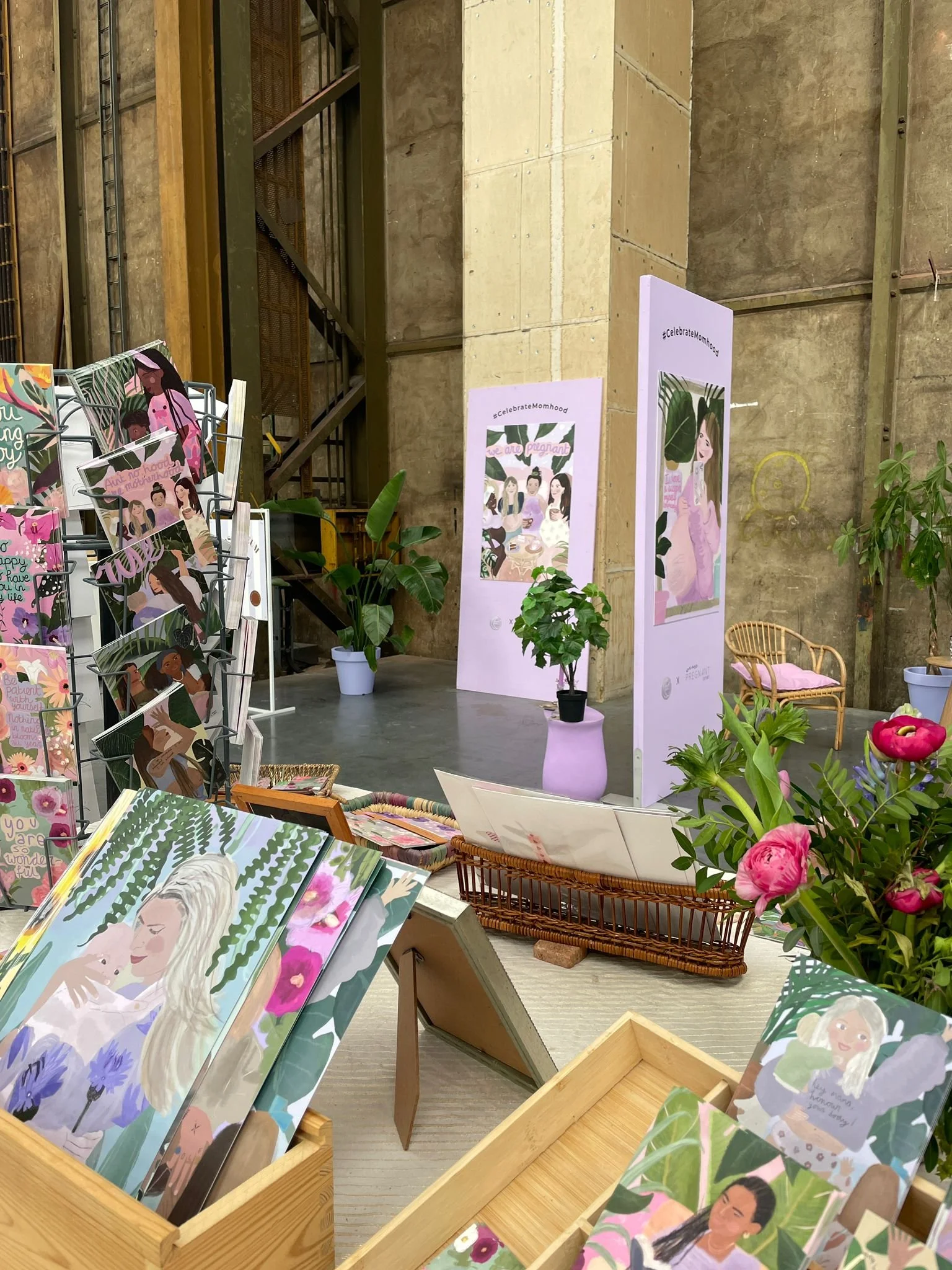 Display of illustrated greeting cards with diverse women, potted plants, and decorative banners in an indoor setting with concrete walls and industrial elements.