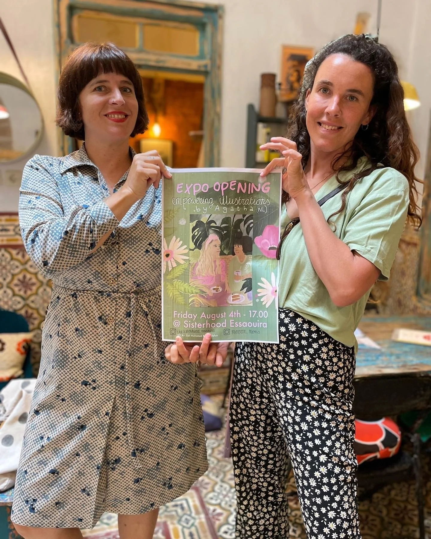 Two women holding a colorful flyer for an expo opening. The flyer features an illustration of two women surrounded by tropical plants and flowers, with event details and social media handles at the bottom. The background shows a cozy indoor space wit