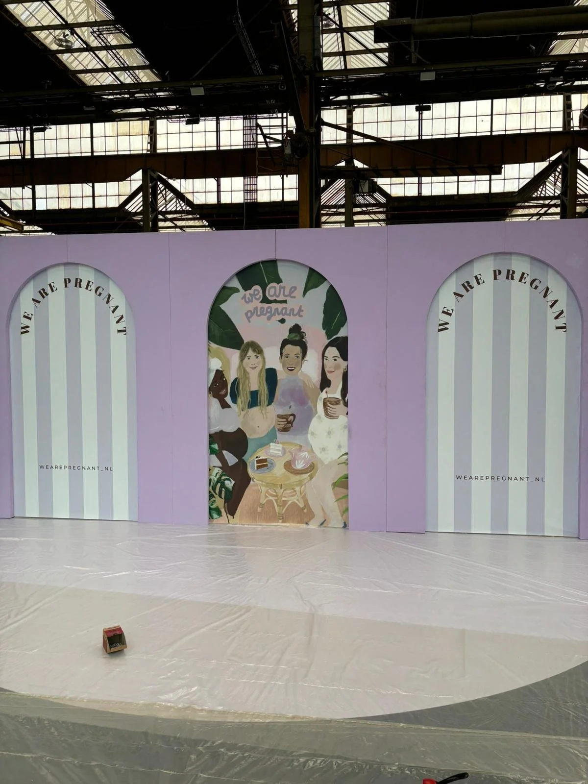 A large indoor space with a high industrial ceiling. A purple backdrop features a mural of three women drinking coffee, with the words 'we are pregnant' written above them. The backdrop repeats 'we are pregnant' on both sides, with the website 'weare