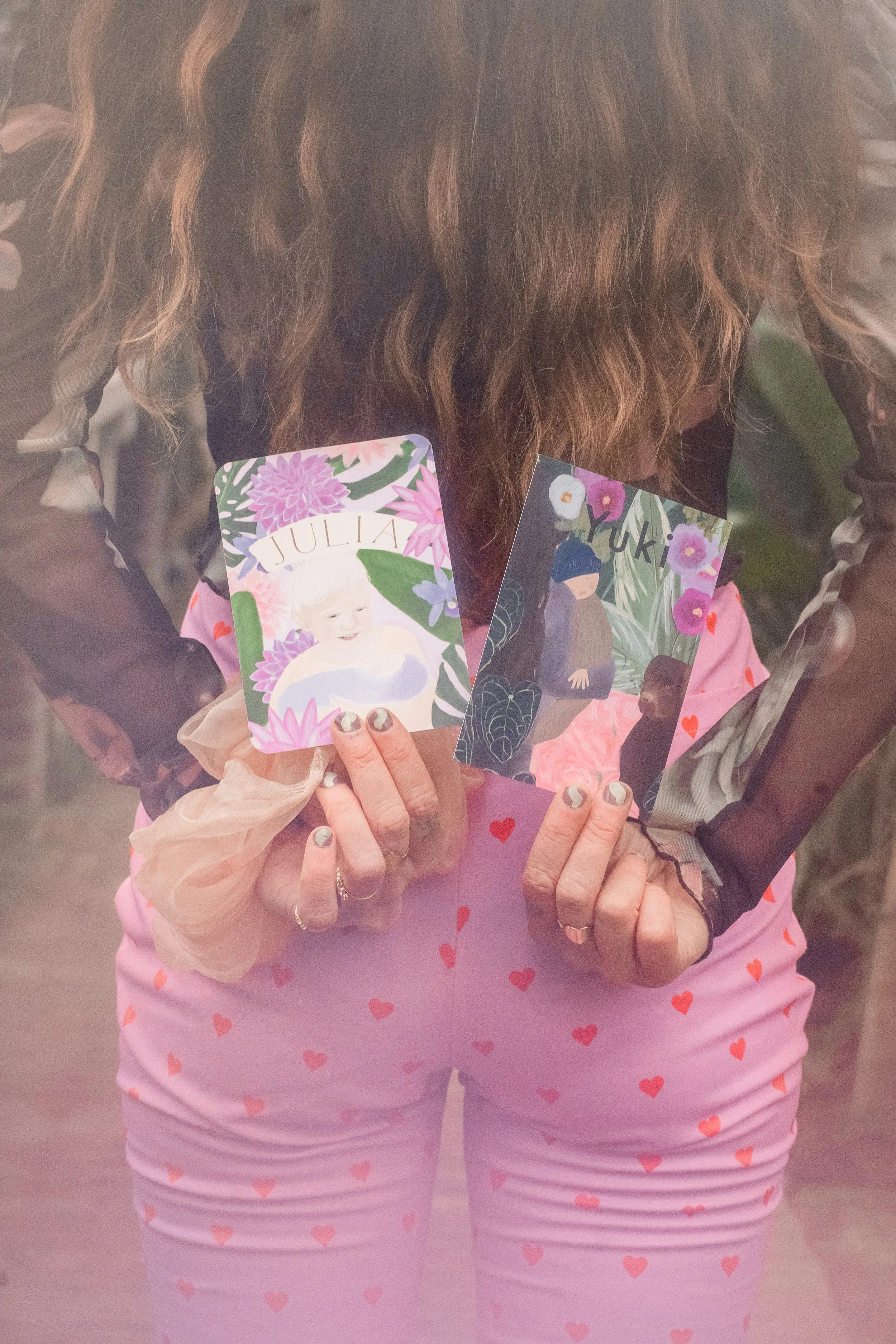 Vrouwelijke illustrator met lang haar houdt twee kleurrijke geboortekaartjes vast, ontworpen met aandacht voor persoonlijke verhaal van het gezin met veel planten en bloemen