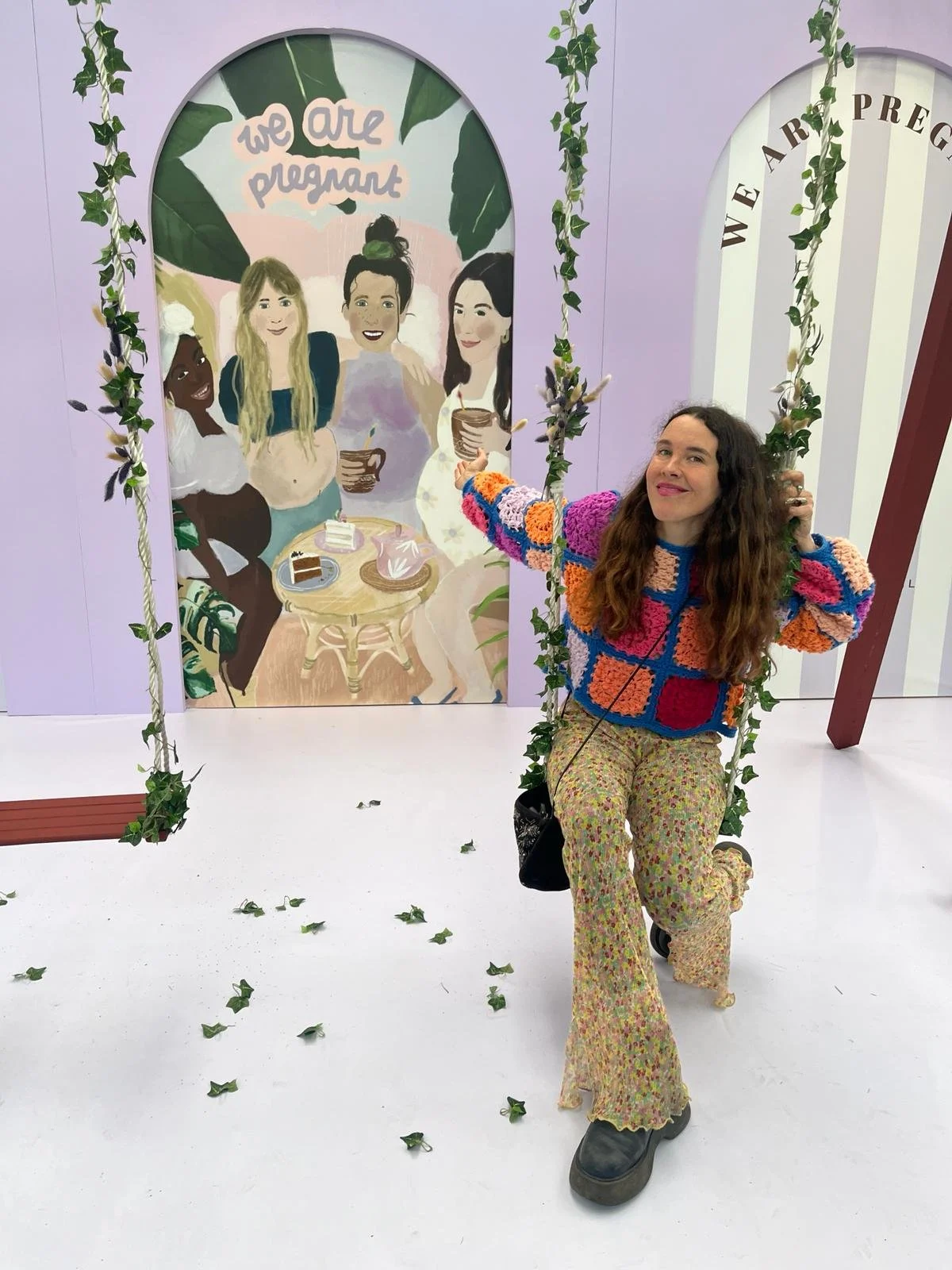 A woman with long, curly hair sitting on a swing decorated with green leaves, in front of a colorful illustrated backdrop featuring diverse women, some holding drinks, with the message "we are pregnant" in a playful font. The swing is part of an indo