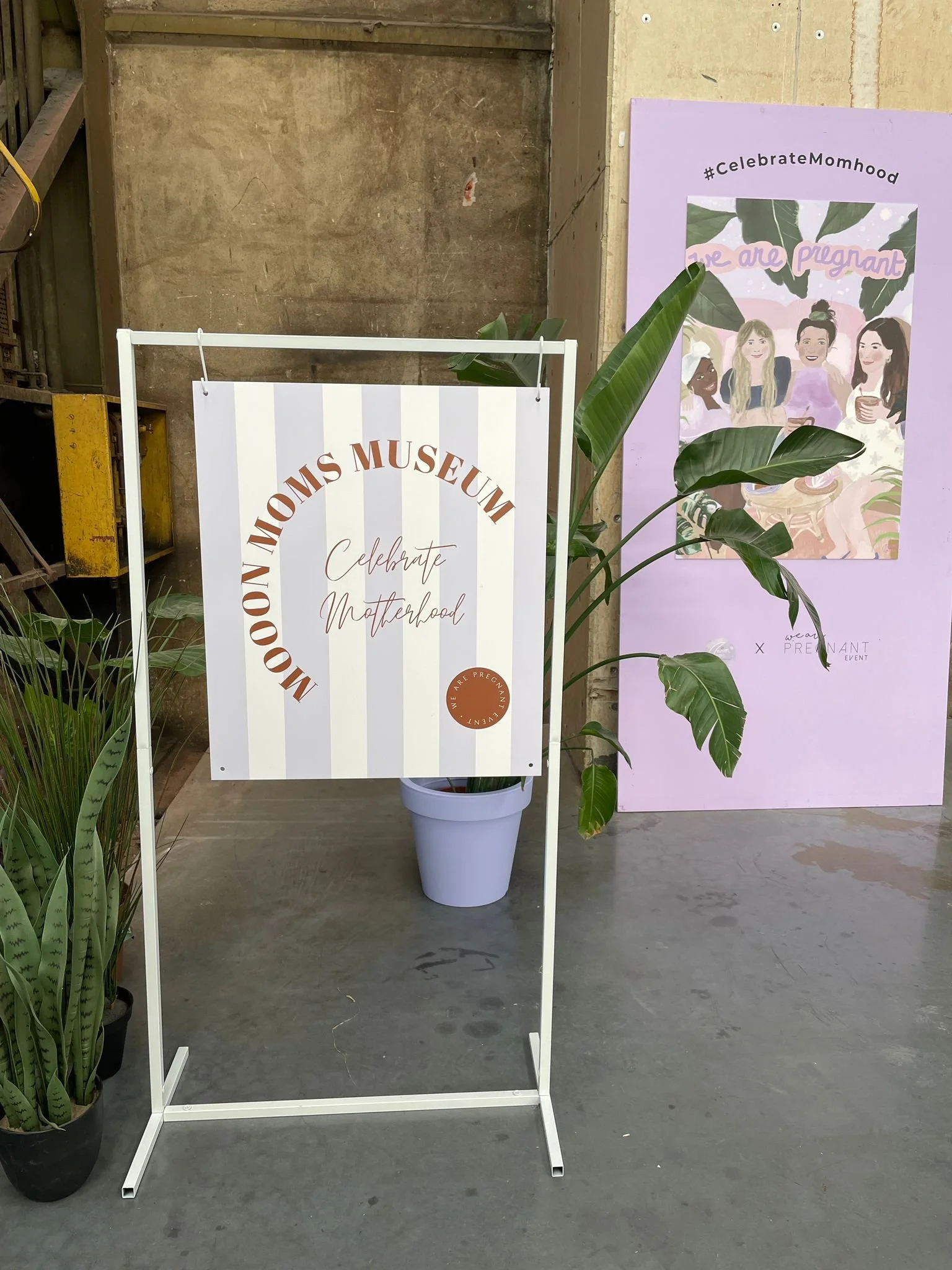 Empty clothing rack with a sign that reads 'Mom's Museum, Celebrate Motherhood,' a large leafy potted plant in front of a purple display poster with illustrations of women, and some potted plants on the left side.