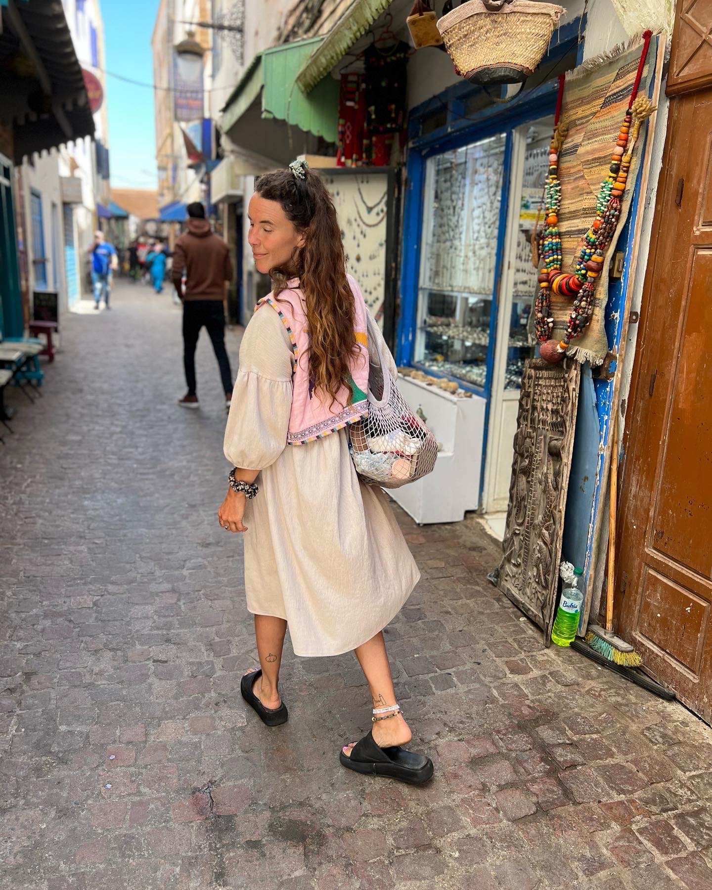 A woman with long wavy brown hair wearing a beige dress, black sandals, and layered ankle jewelry, standing on a cobblestone street in a colorful marketplace with jewelry displays and a green bottle of water at her feet.