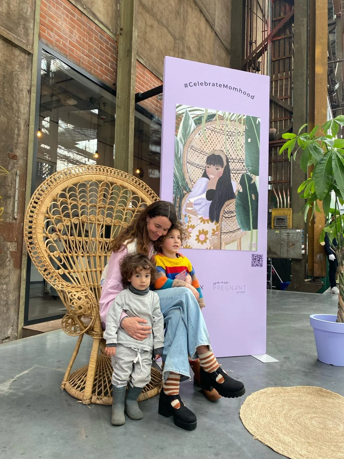 A woman with long brown hair sitting on a wicker peacock chair with two children on her lap and standing next to her, indoors with a large purple sign in the background that has an illustration of a woman in a hat and the hashtag #CelebrateMomhood.