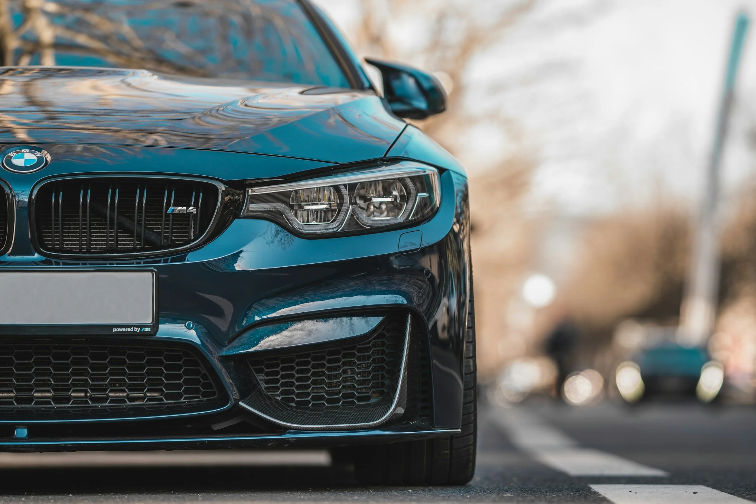Close-up of a blue BMW M4 parked on the street, focusing on the front left side, including the grille, headlight, and part of the bumper.