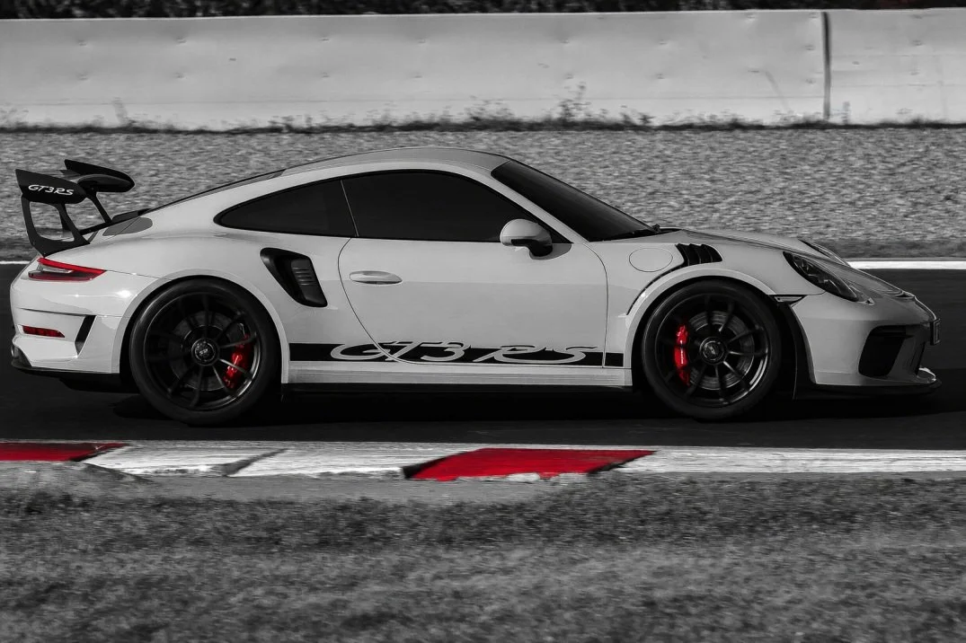 A white Porsche 911 GT3 RS on a racetrack with red brake calipers, black wheels, and a large rear wing, in a mostly black and white image with selective red accents.