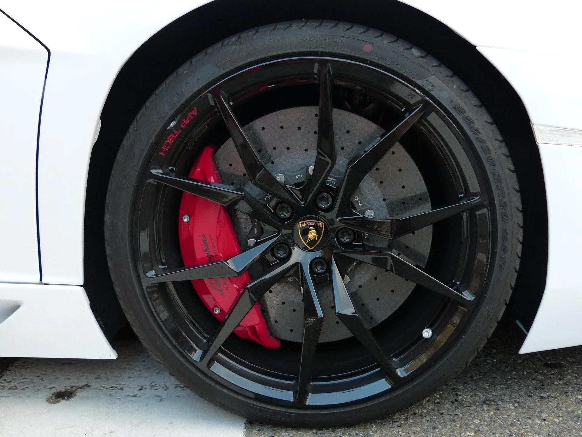 Close-up of a white luxury sports car wheel with a black rim, red brake caliper, and drilled brake disc, featuring an Lamborghini logo in the center.