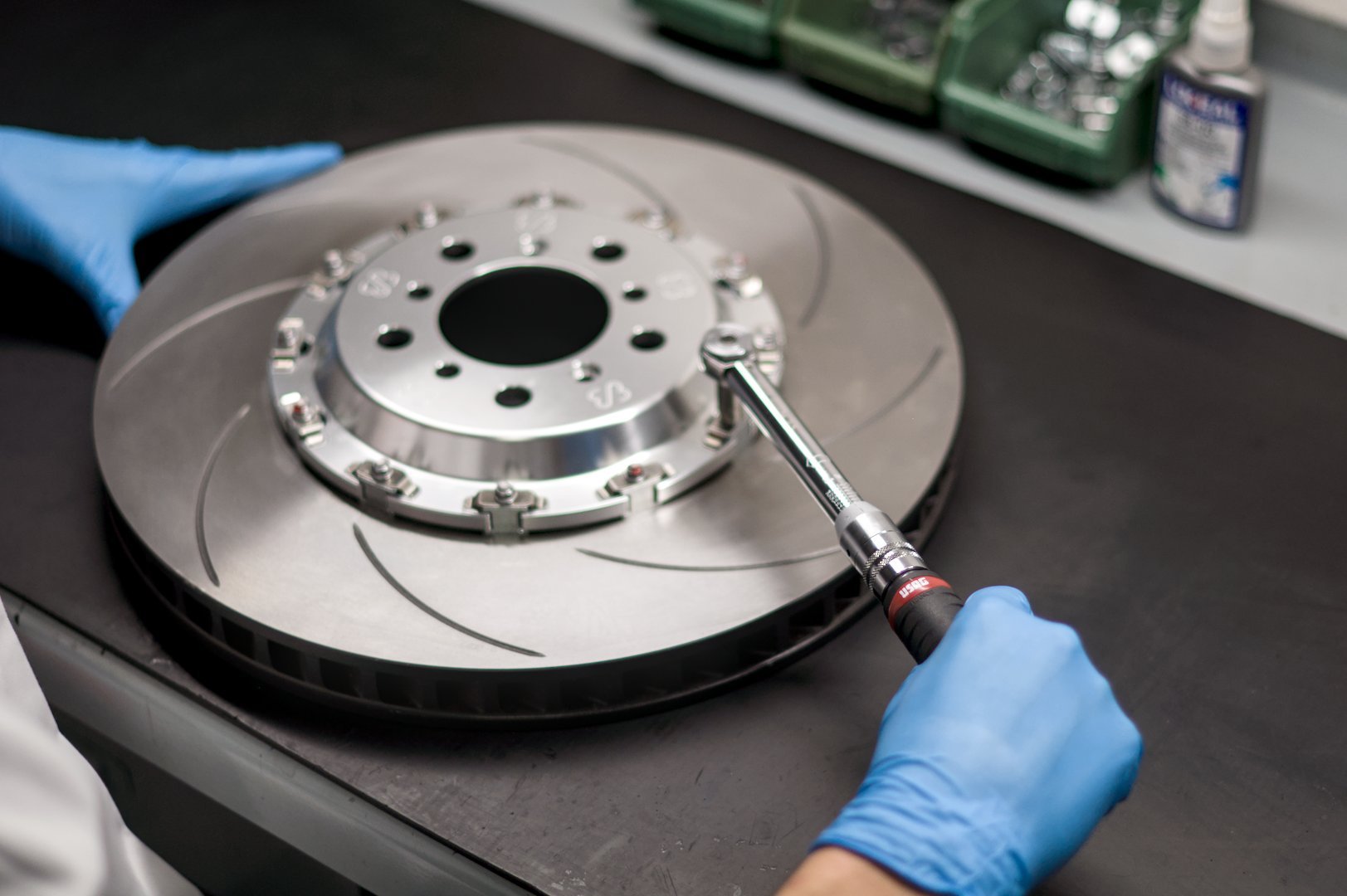 Person wearing blue gloves working on a large metal brake rotor with a torque wrench on a workbench in a workshop