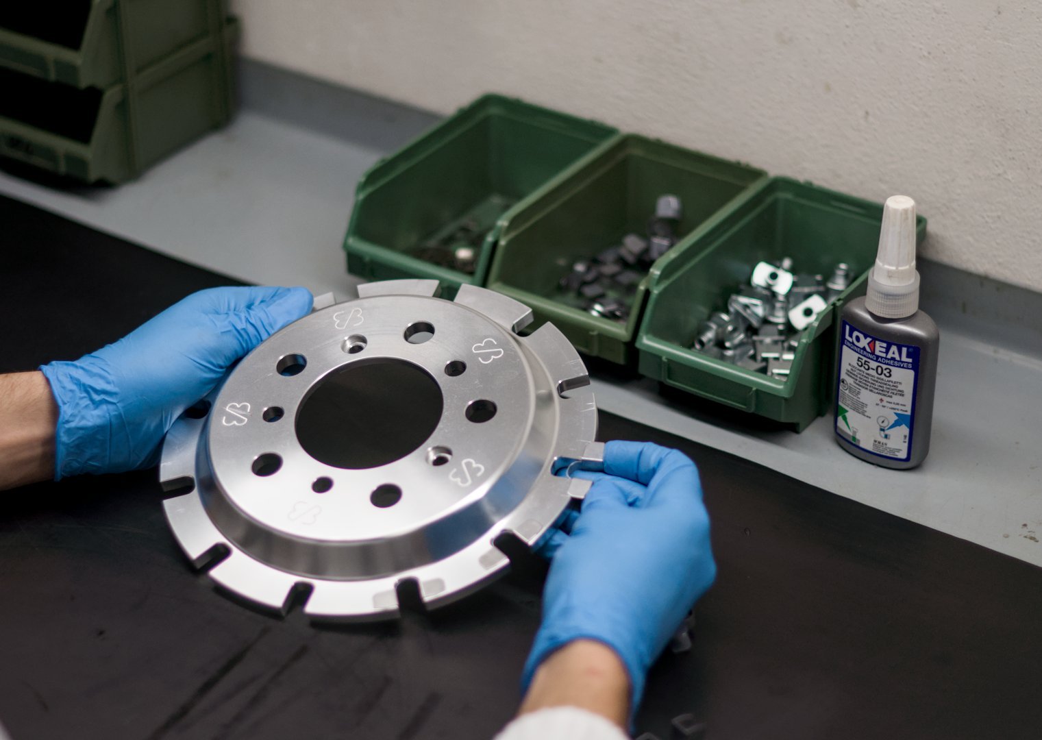Person wearing blue gloves assembling a circular metallic part on a black work surface with green containers holding small hardware pieces and a bottle of Loctite adhesive in the background.