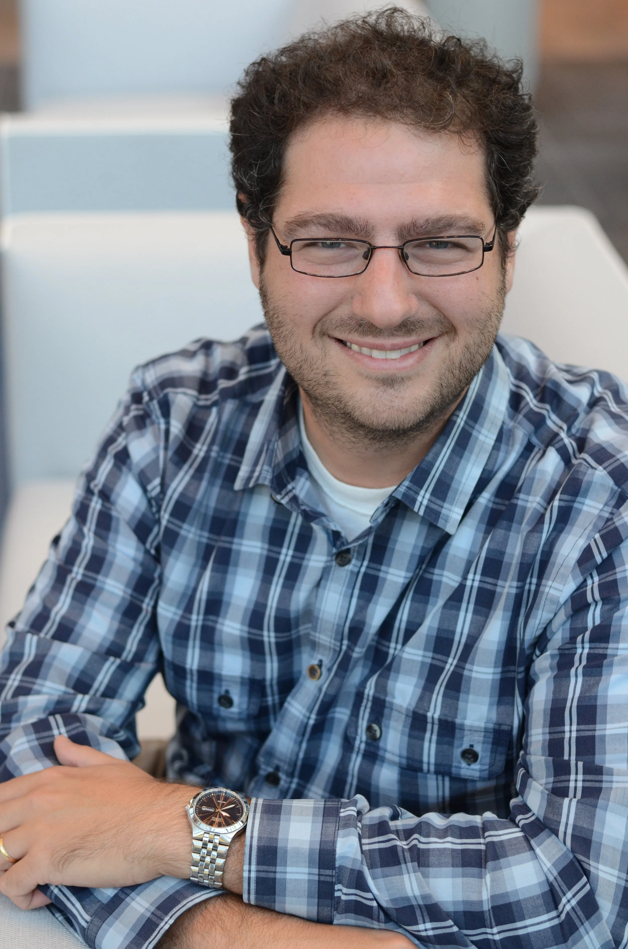A smiling man with glasses and a beard wearing a blue plaid shirt. He's sitting at a table with his arms crossed, wearing a watch. The background shows modern office or lounge seating.