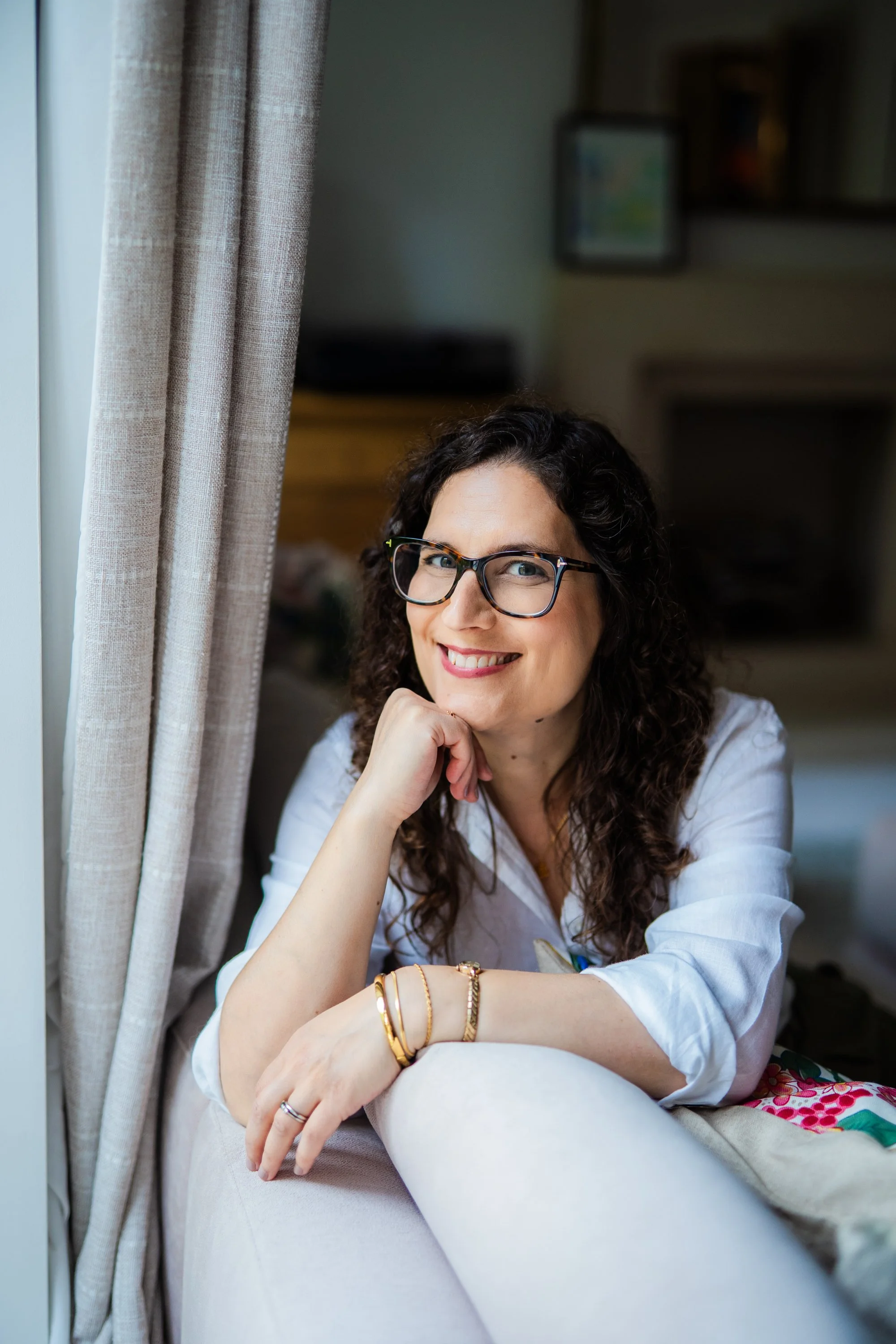 A woman with curly dark hair and glasses smiling and resting her chin on her hand, sitting near a window with curtains.