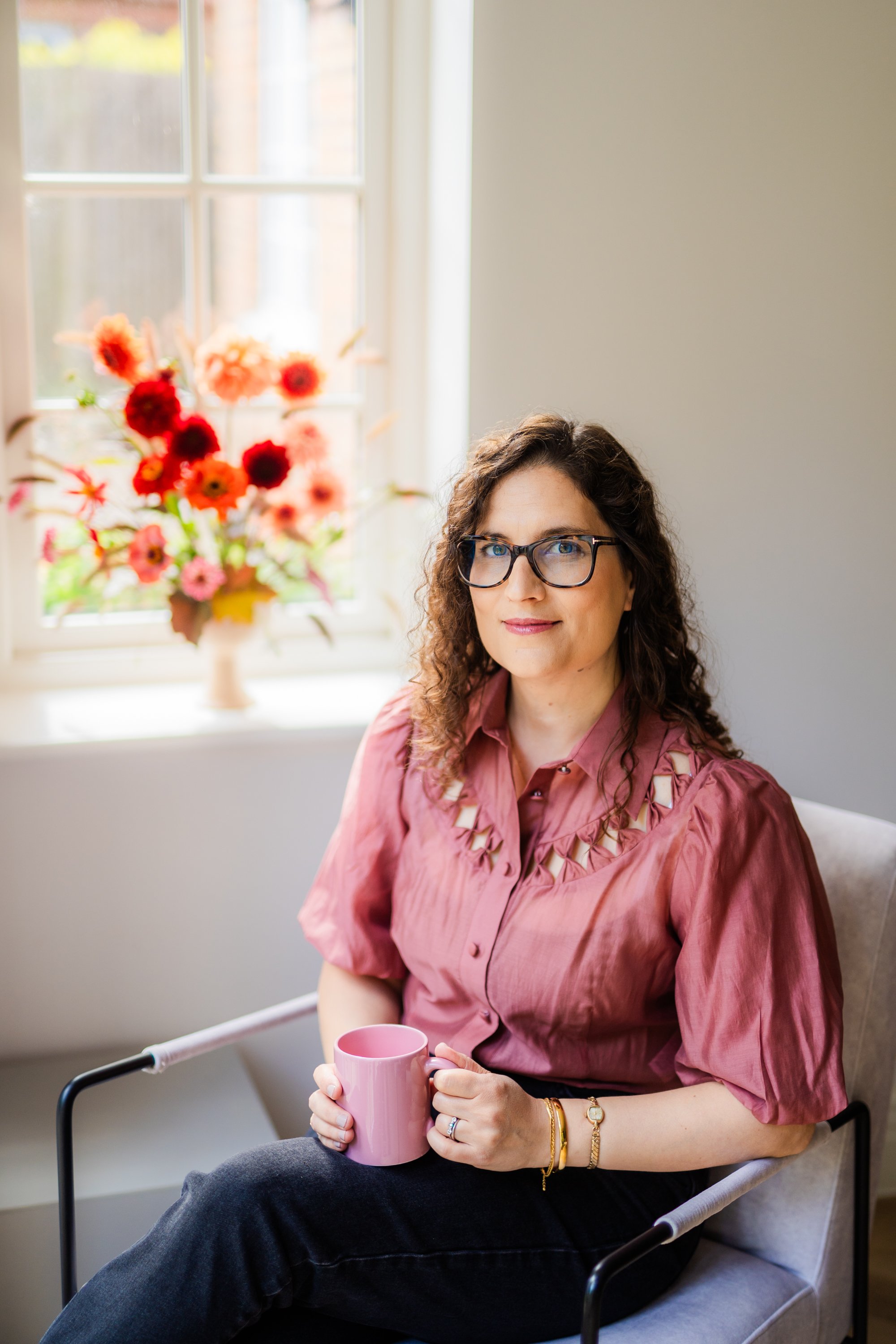 A woman with curly brown hair and glasses sitting in a chair, holding a pink mug in her right hand. She is wearing a pink blouse with cutout details on the shoulders. There is a window with sunlight and a vase of colorful flowers on the windowsill in the background.