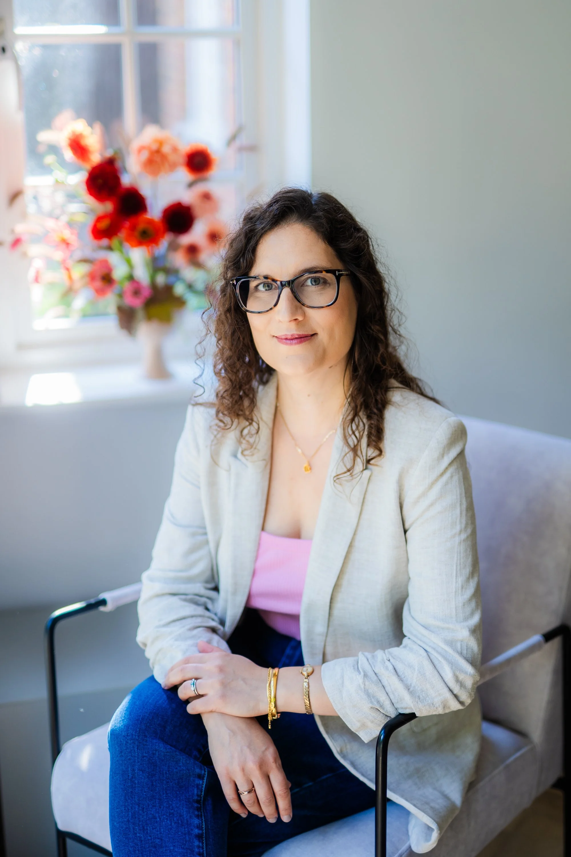 A woman with curly brown hair, glasses, and light skin sitting on a chair in a brightly lit room with a vase of colorful flowers on the windowsill behind her.