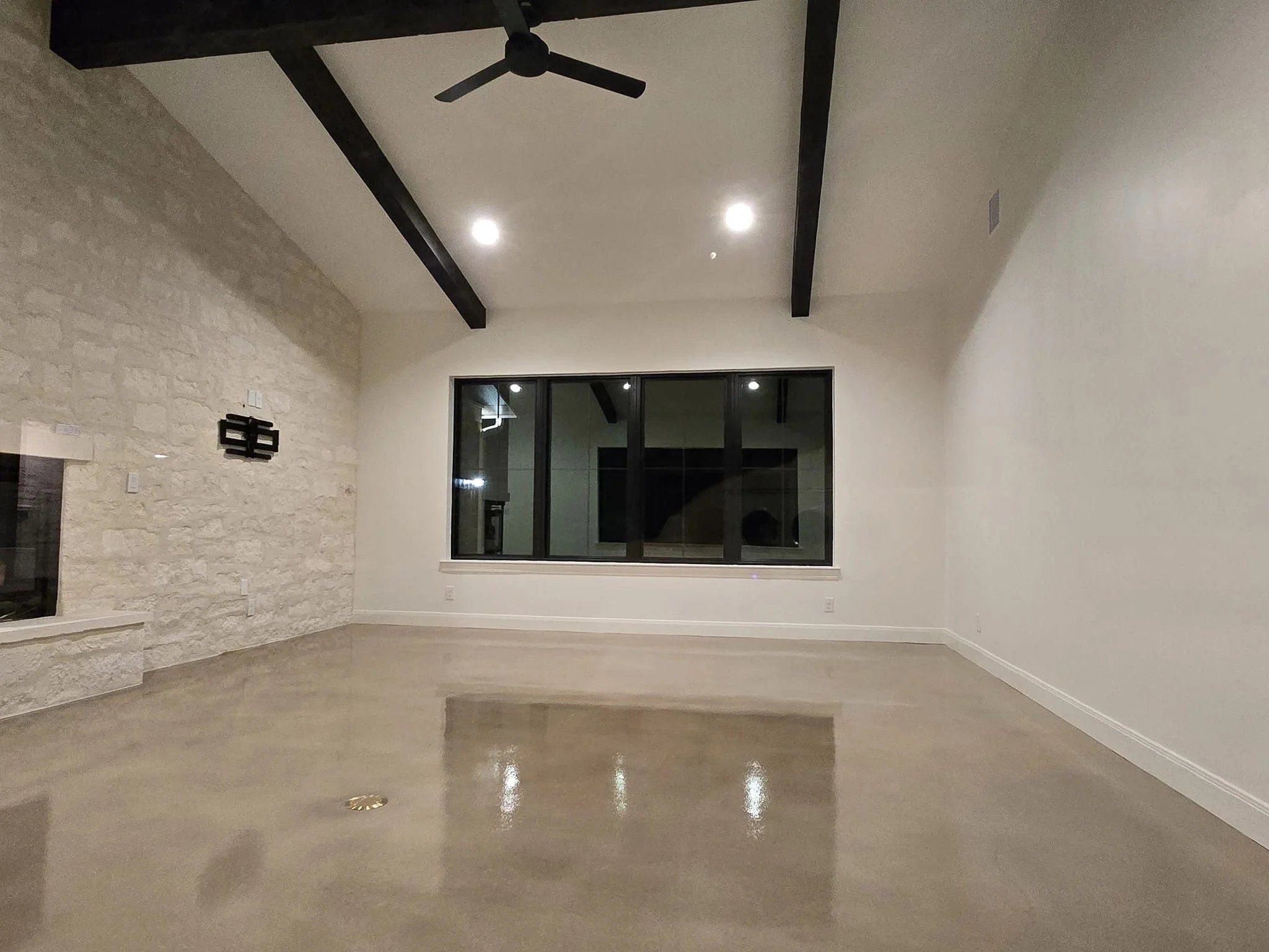 Empty living room with polished concrete floors, white walls, a large window, black ceiling beams, a ceiling fan, and a fireplace with a stone wall.