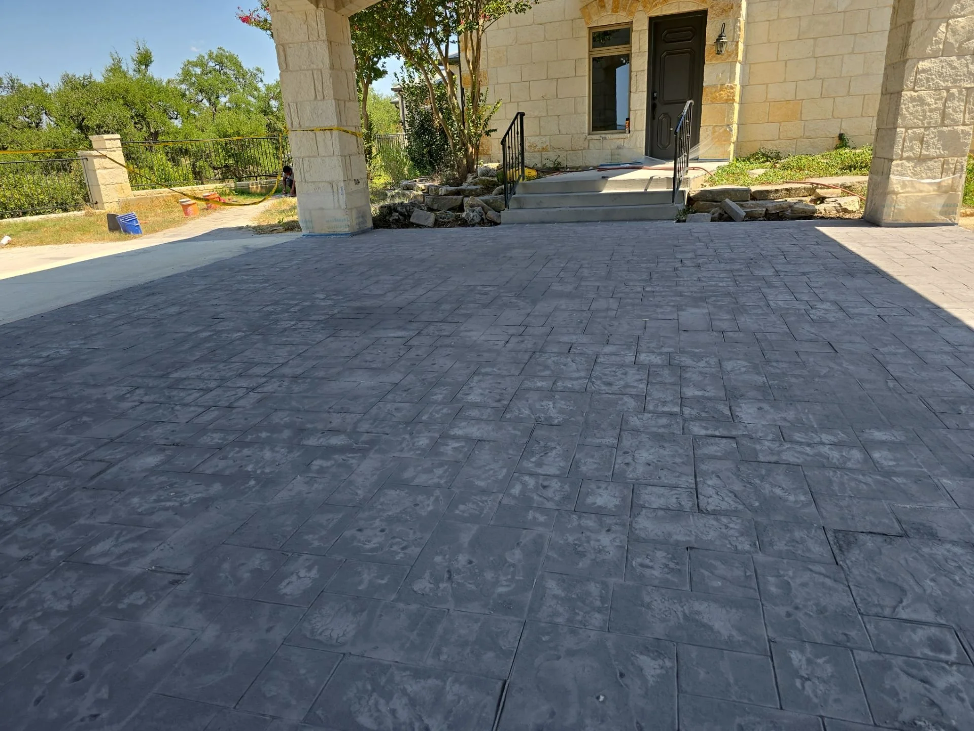Newly paved concrete driveway leading to a house with a front porch, steps, and stone columns, surrounded by trees and yard.