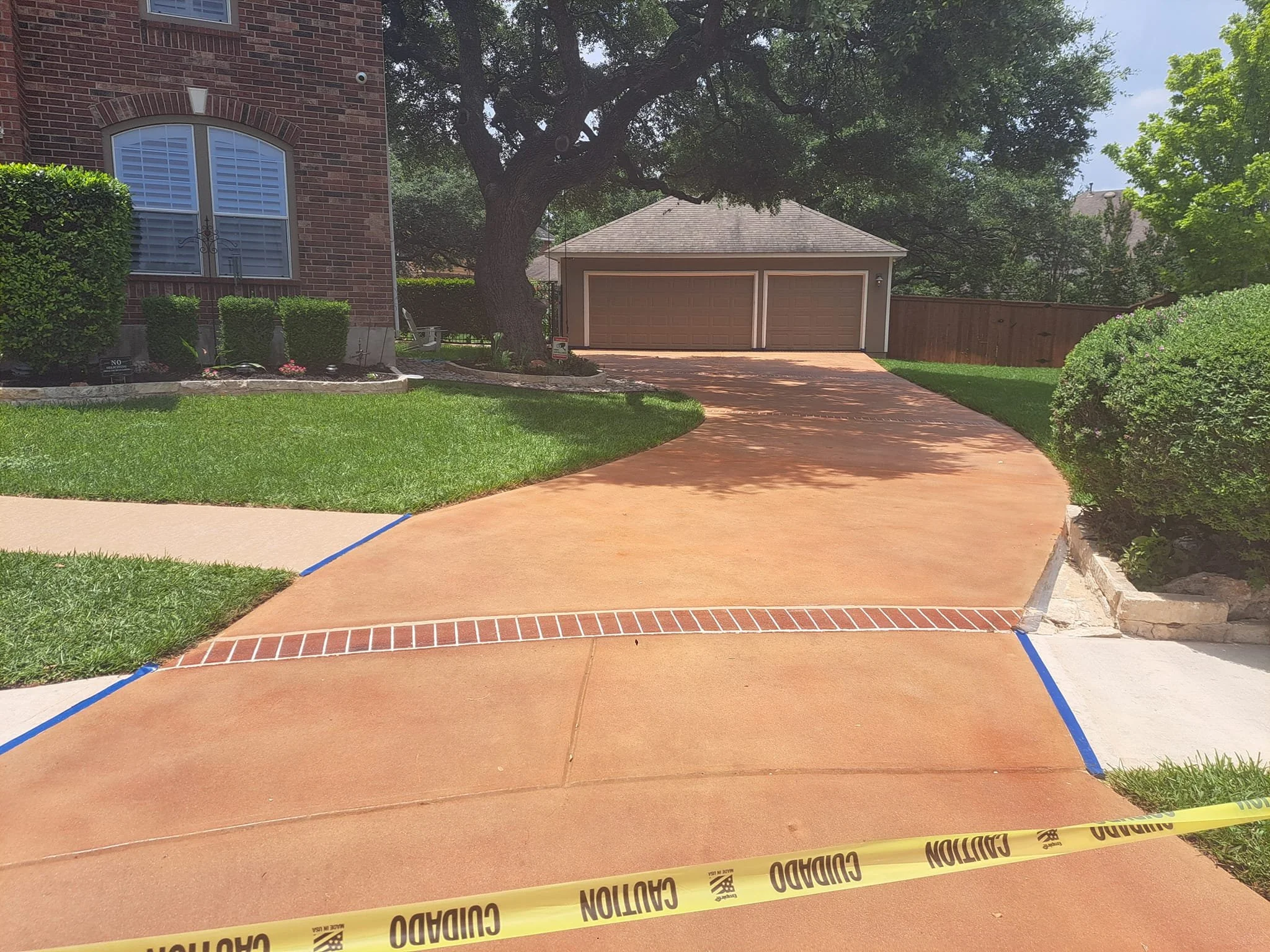 A new concrete driveway leading to a garage, with yellow caution tape across it, part of a residential yard with green grass, bushes, and trees.