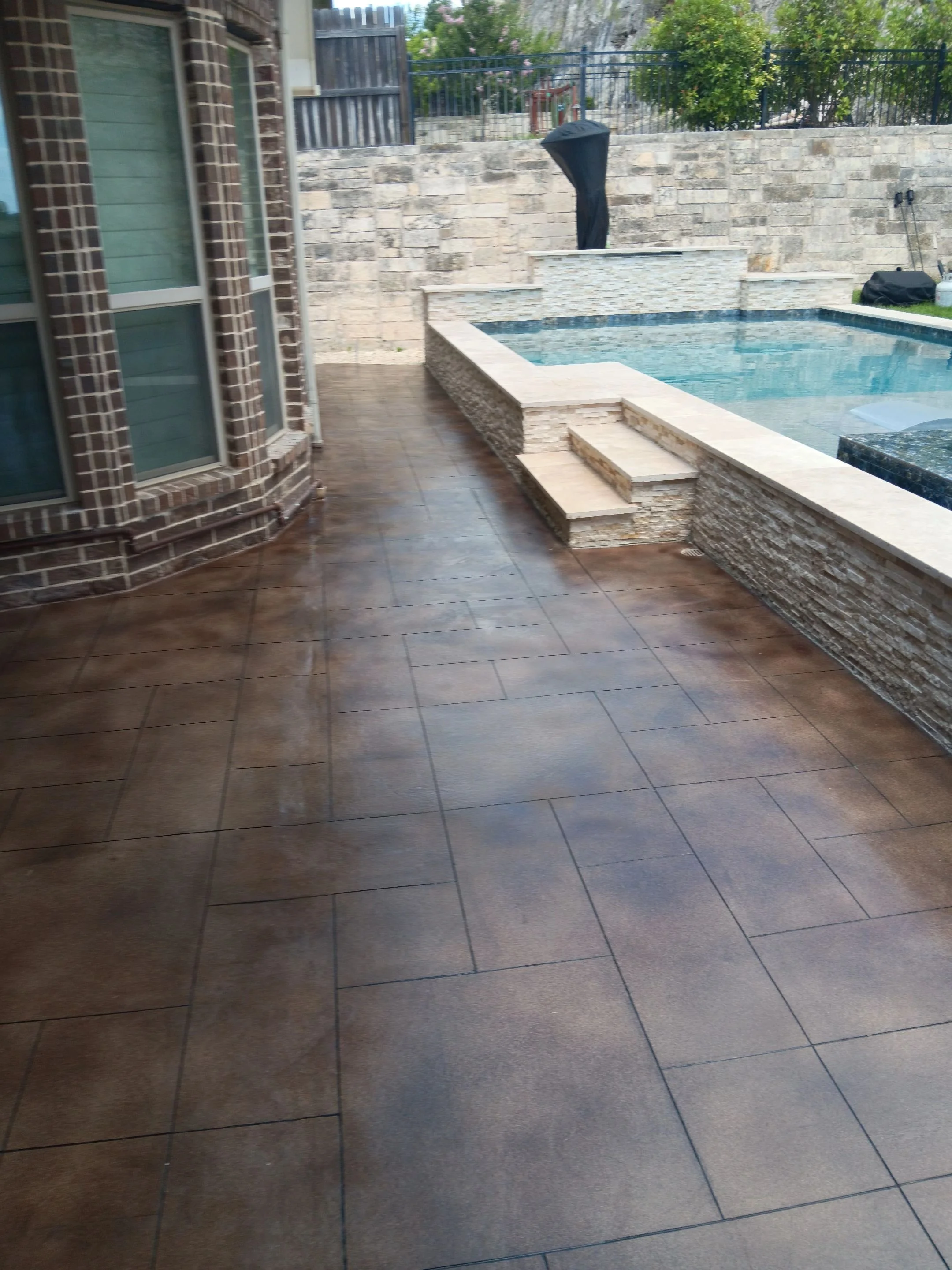 Poolside patio with wet, brown tile flooring, brick exterior corner, and a swimming pool with a stone edge and steps, enclosed by a stone wall with trees and fence in the background.