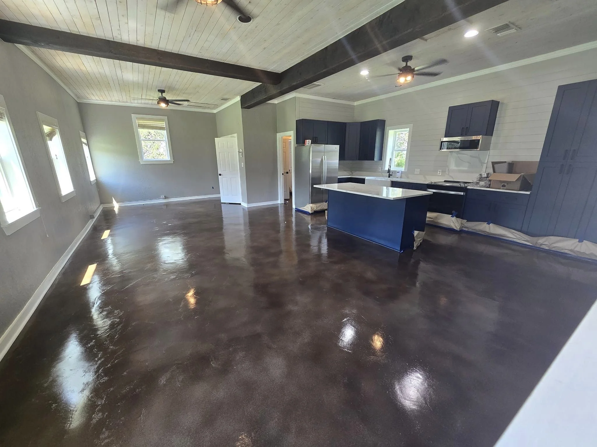 Open-concept living space with dark glossy stained concrete floors, light gray walls, a vaulted ceiling with wood beams and ceiling fans, and a kitchen with dark blue cabinets, a white countertop island, stainless steel appliances, and multiple windo