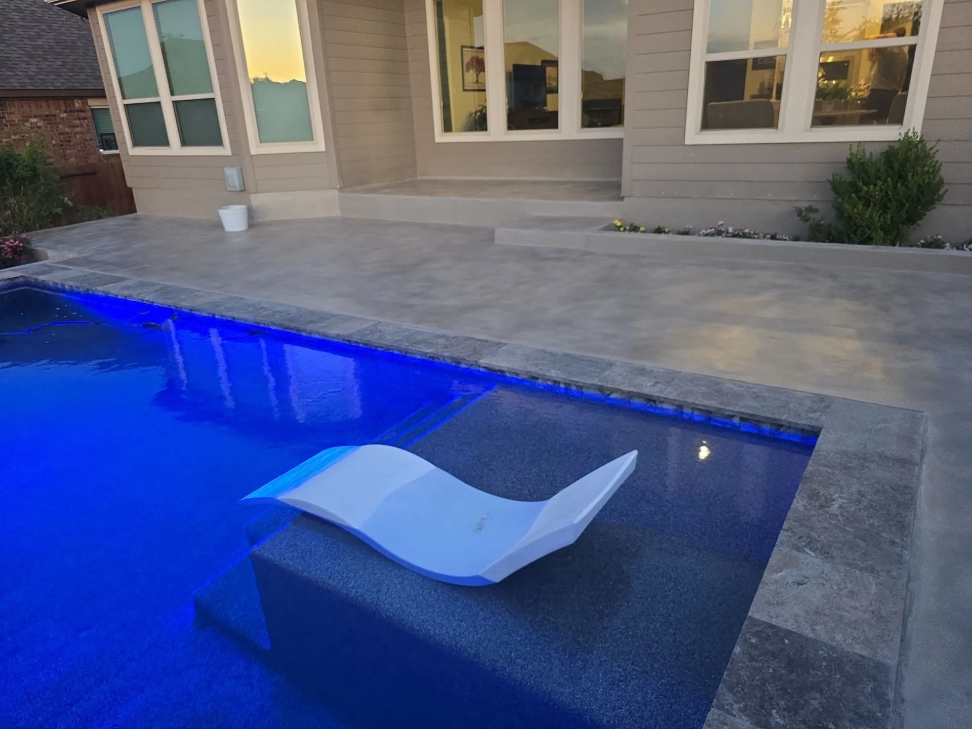 Swimming pool with a white lounge chair floating in the water, house exterior with large windows and a deck in the background, evening lighting.
