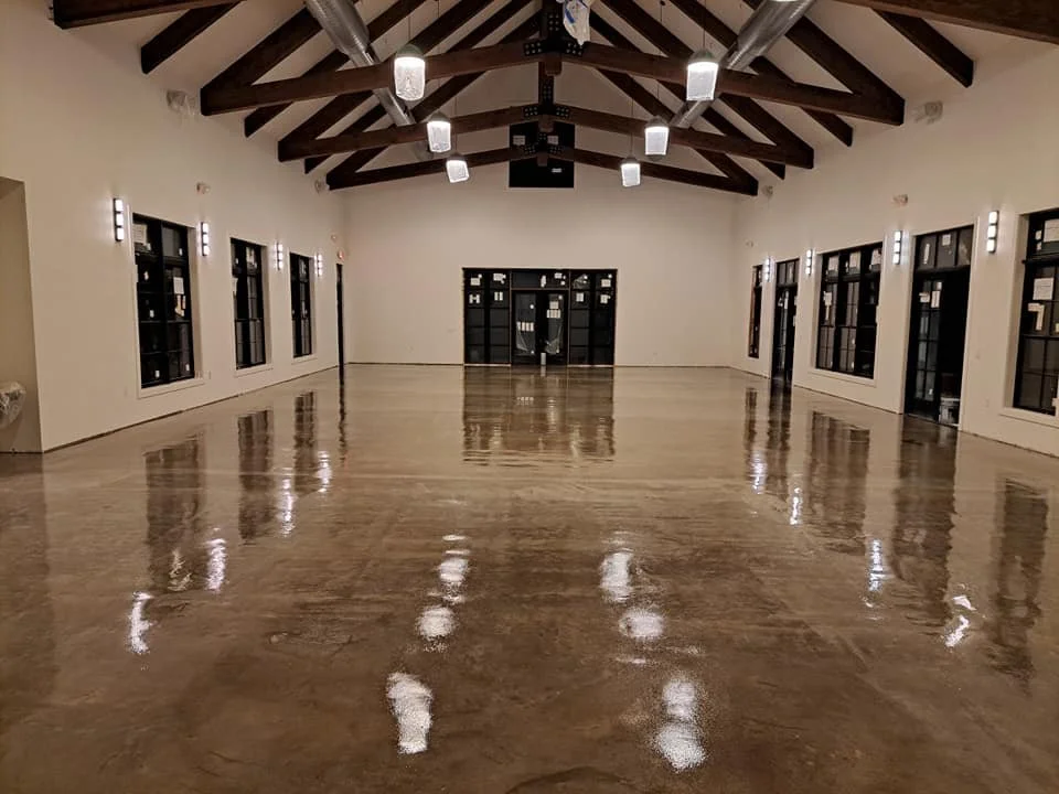 Empty spacious room with polished wooden floor, white walls, multiple windows along sides, and a vaulted ceiling with exposed dark wooden beams and hanging lights.
