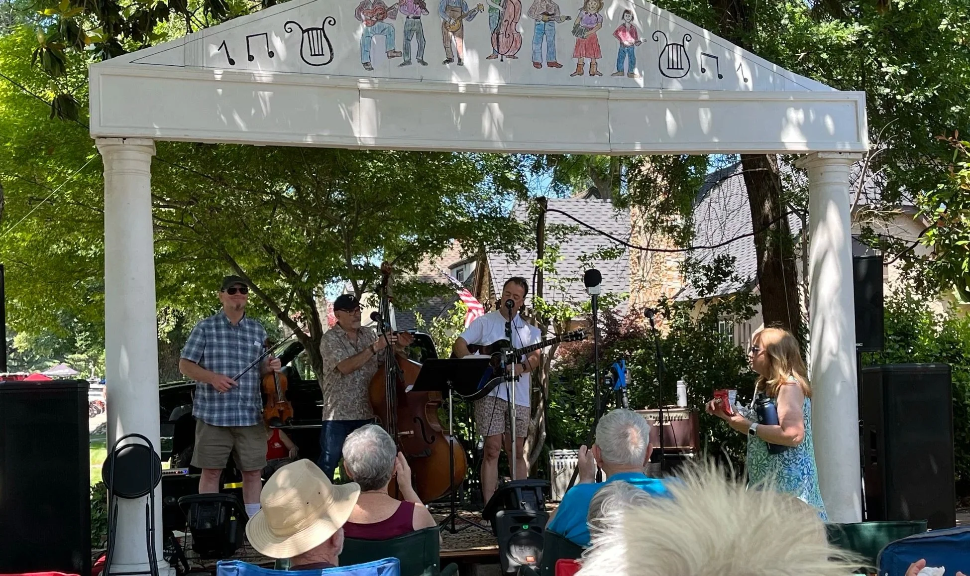 A band performing live on an outdoor stage with a white classical style arch and columns, surrounded by green trees. The band includes musicians playing string instruments and a person singing and playing guitar. Audience members are seated, some wearing hats, enjoying the performance.
