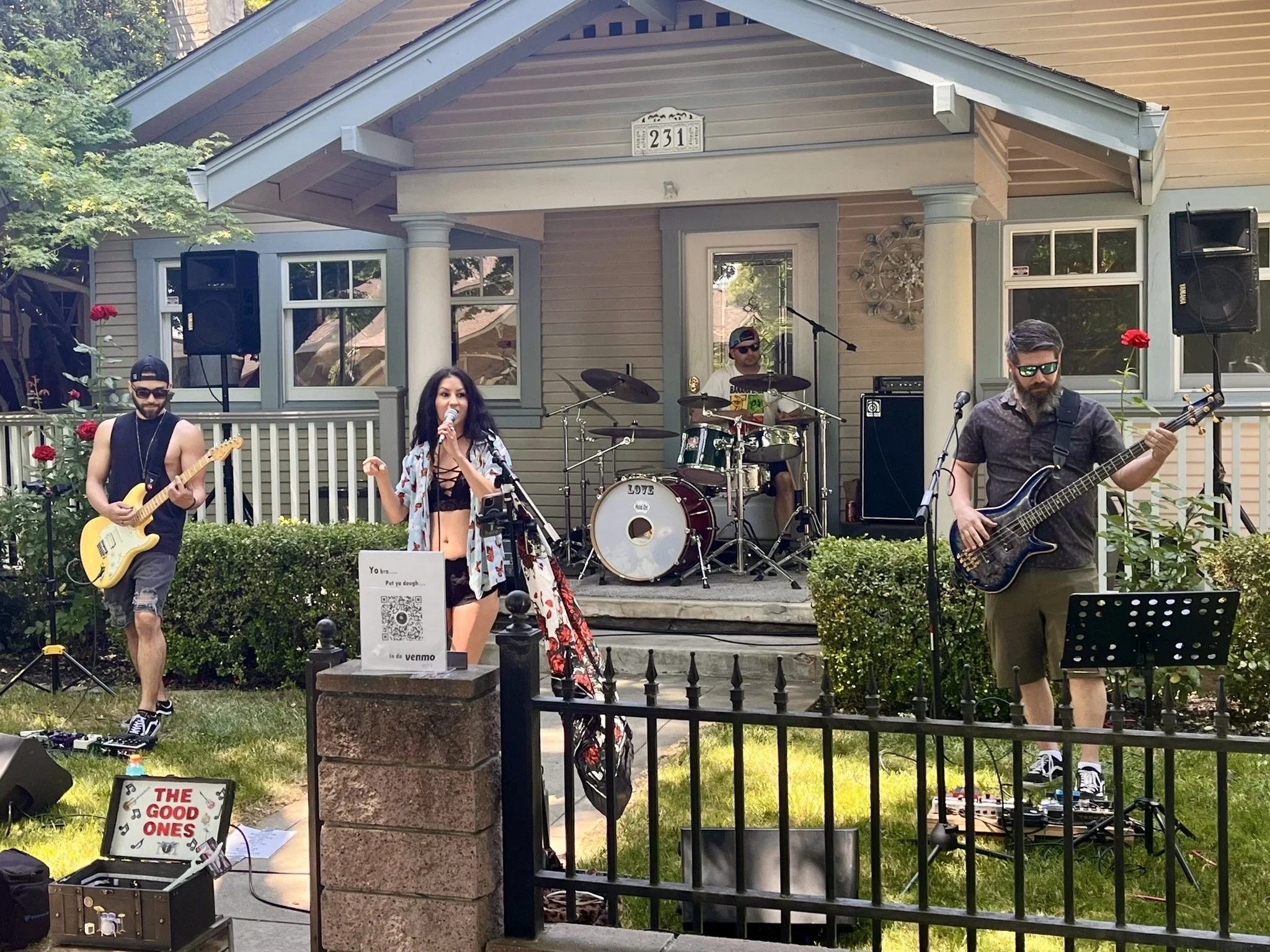 A four-member band performs outdoors on a small stage in front of a house, with two male guitarists, a female vocalist, and a male drummer. The setting is a garden with greenery and roses, during daytime.