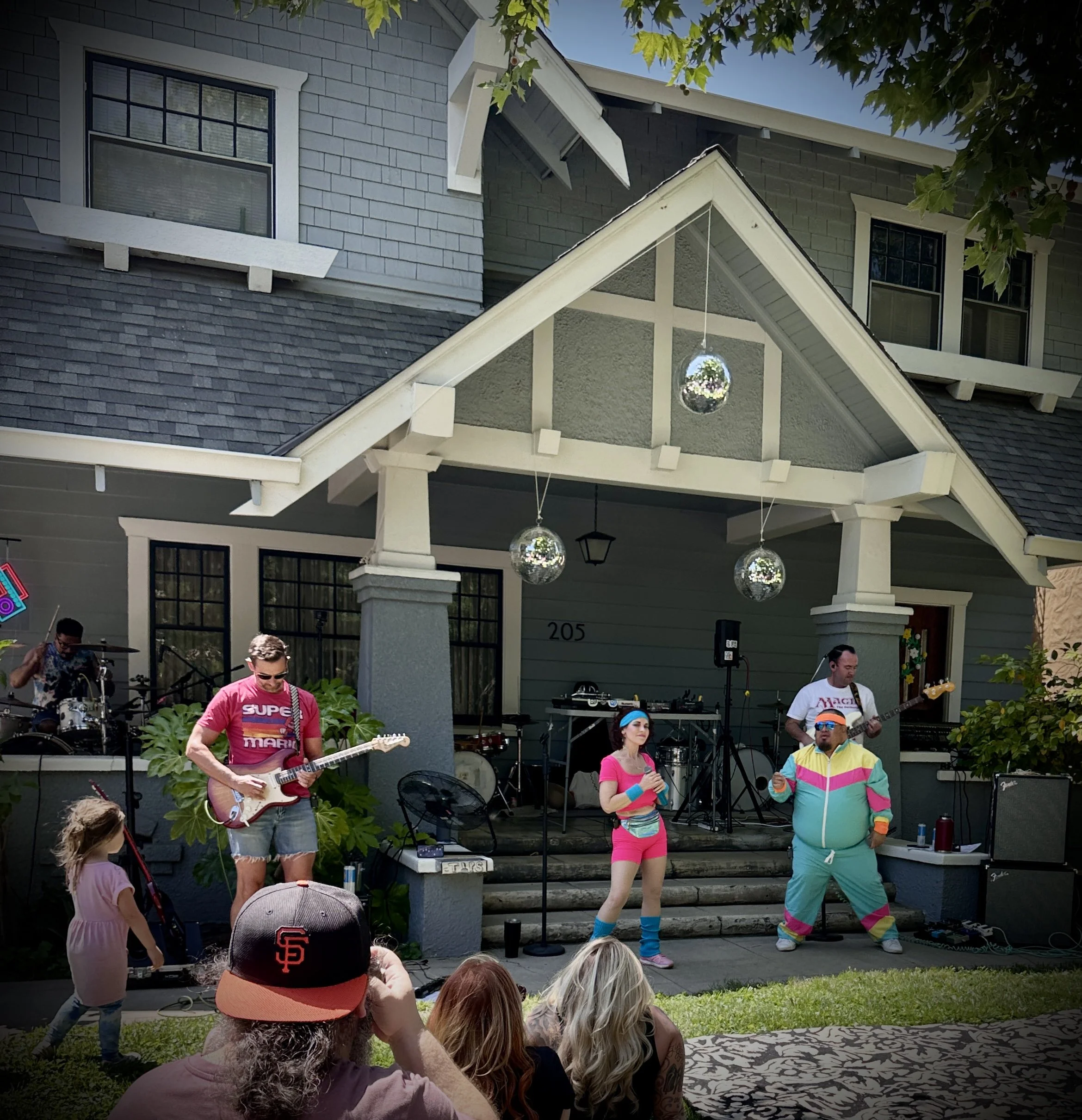 A live outdoor musical performance on a porch of a house, with a band playing instruments and two singers, one in pink and the other in colorful retro tracksuit, surrounded by audience members including children and adults.
