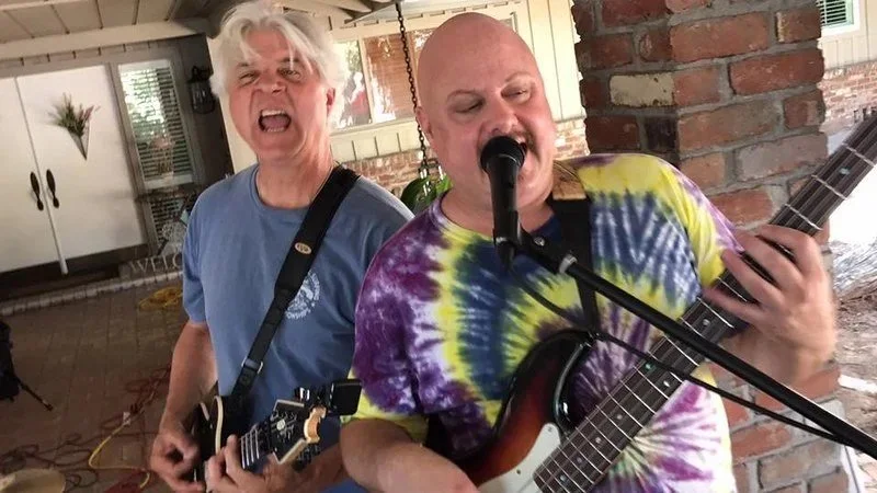 Two men playing guitars and singing passionately, one in a blue shirt and the other in a tie-dye shirt, inside a brick-walled room.