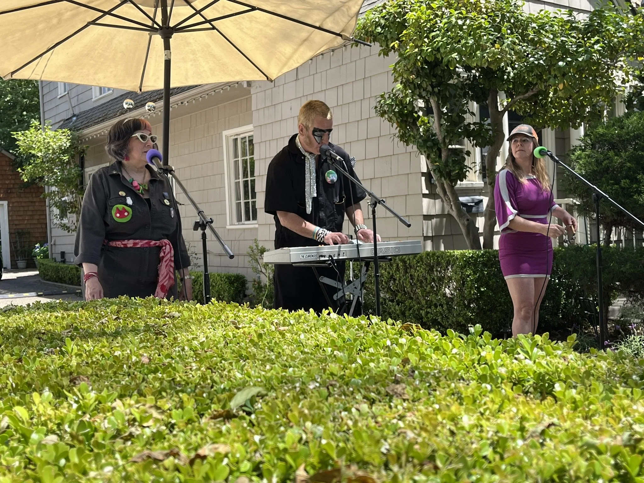 Three musicians perform on an outdoor stage. The woman on the left wears white sunglasses, a black jacket with patches, and a red scarf, singing into a purple microphone. The man in the middle plays a keyboard, with face paint, glasses, and a black shirt. The woman on the right wears a purple dress and a cap, singing into a green microphone. They are under a large umbrella, with green bushes and a house in the background.